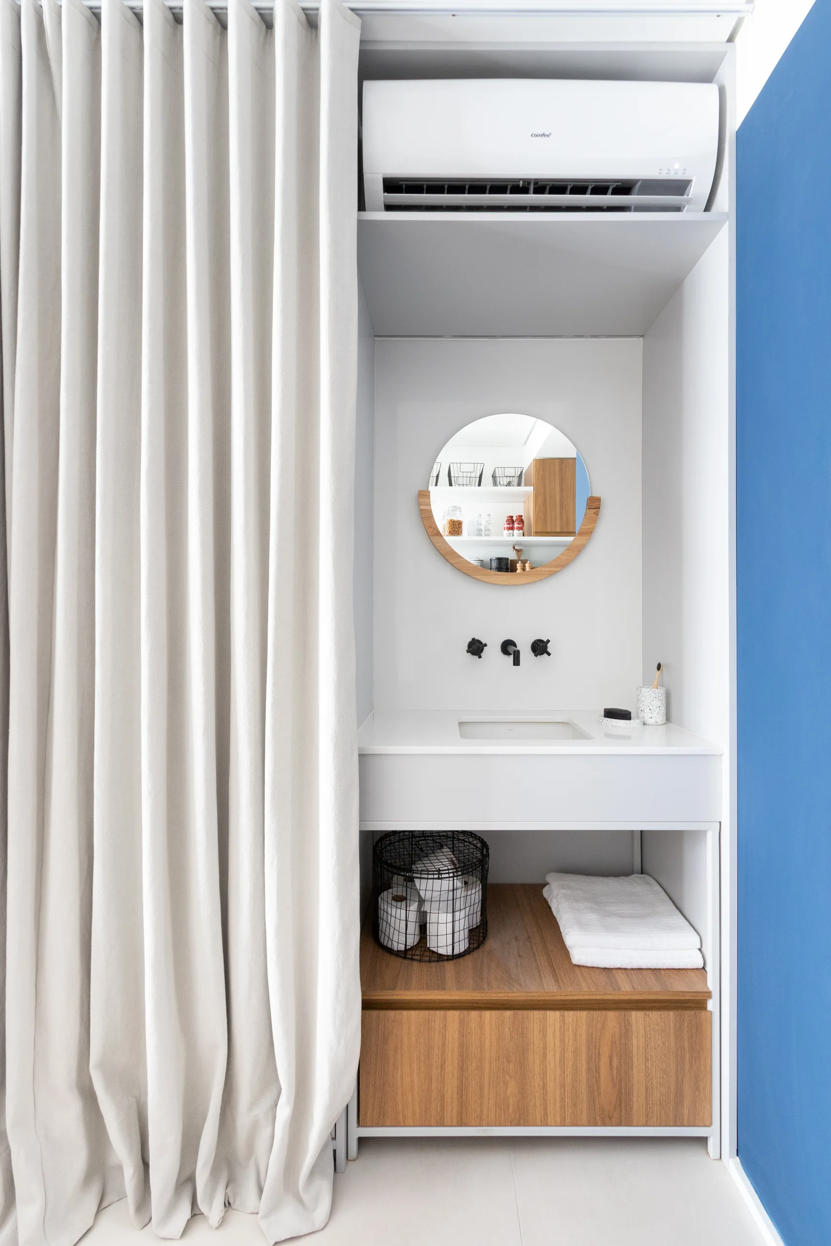A compact white bathroom vanity with a round wooden mirror, a wall-mounted black faucet, lower wood storage shelf, and a light-colored curtain.