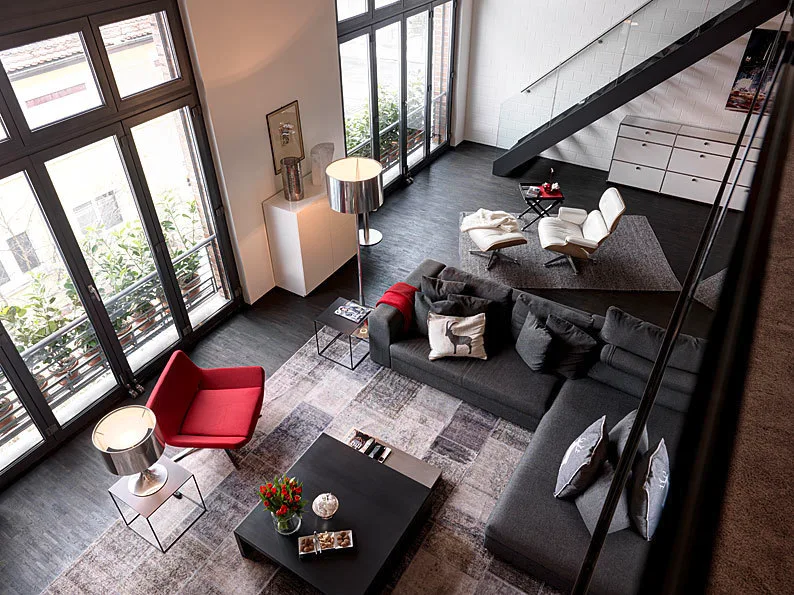 A high-angle view of a contemporary loft living room with a gray sectional sofa, a red accent chair, and large dark-framed windows.