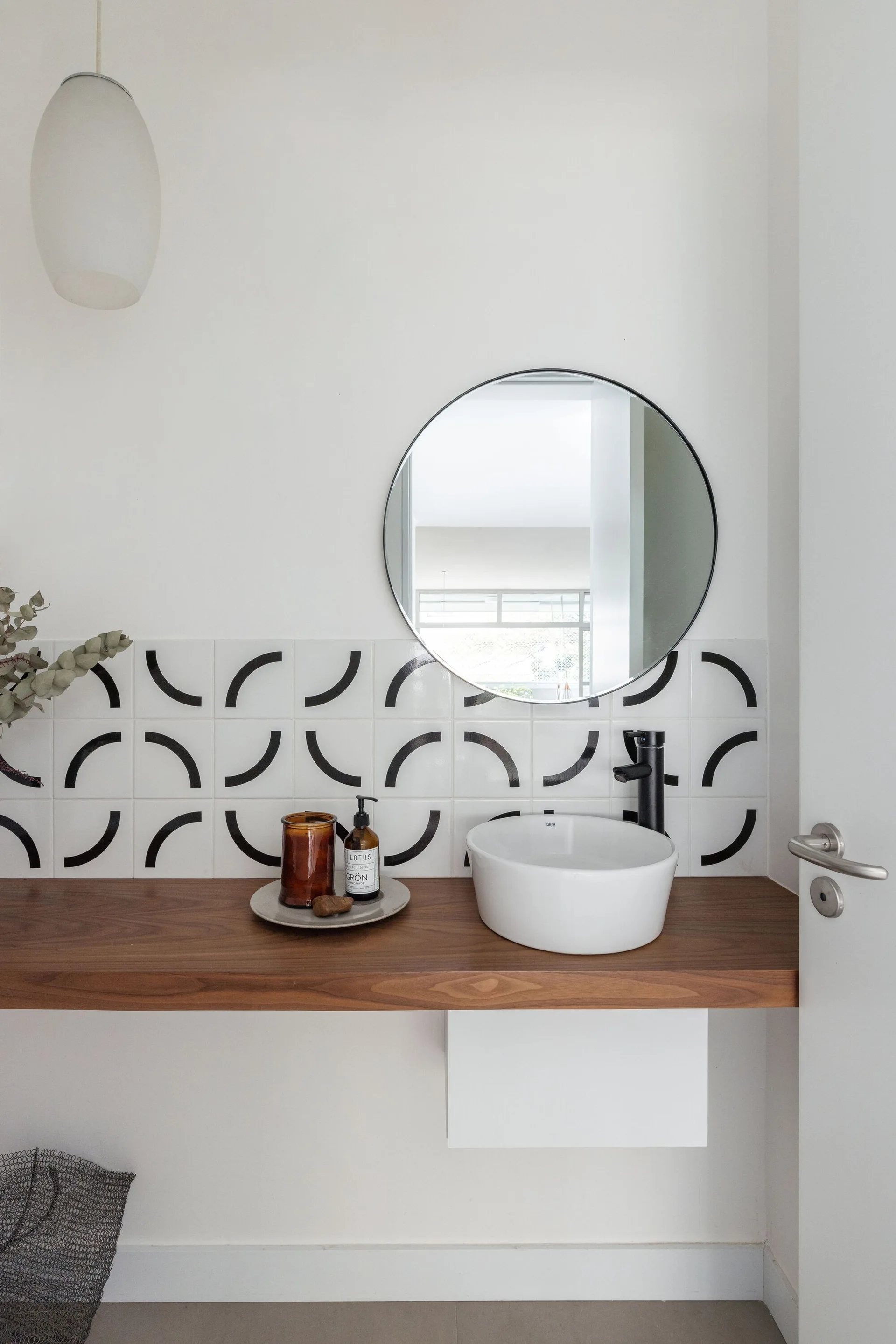 A floating wood vanity features a white vessel sink, black faucet, round mirror, and a geometric backsplash tile pattern of black arcs.