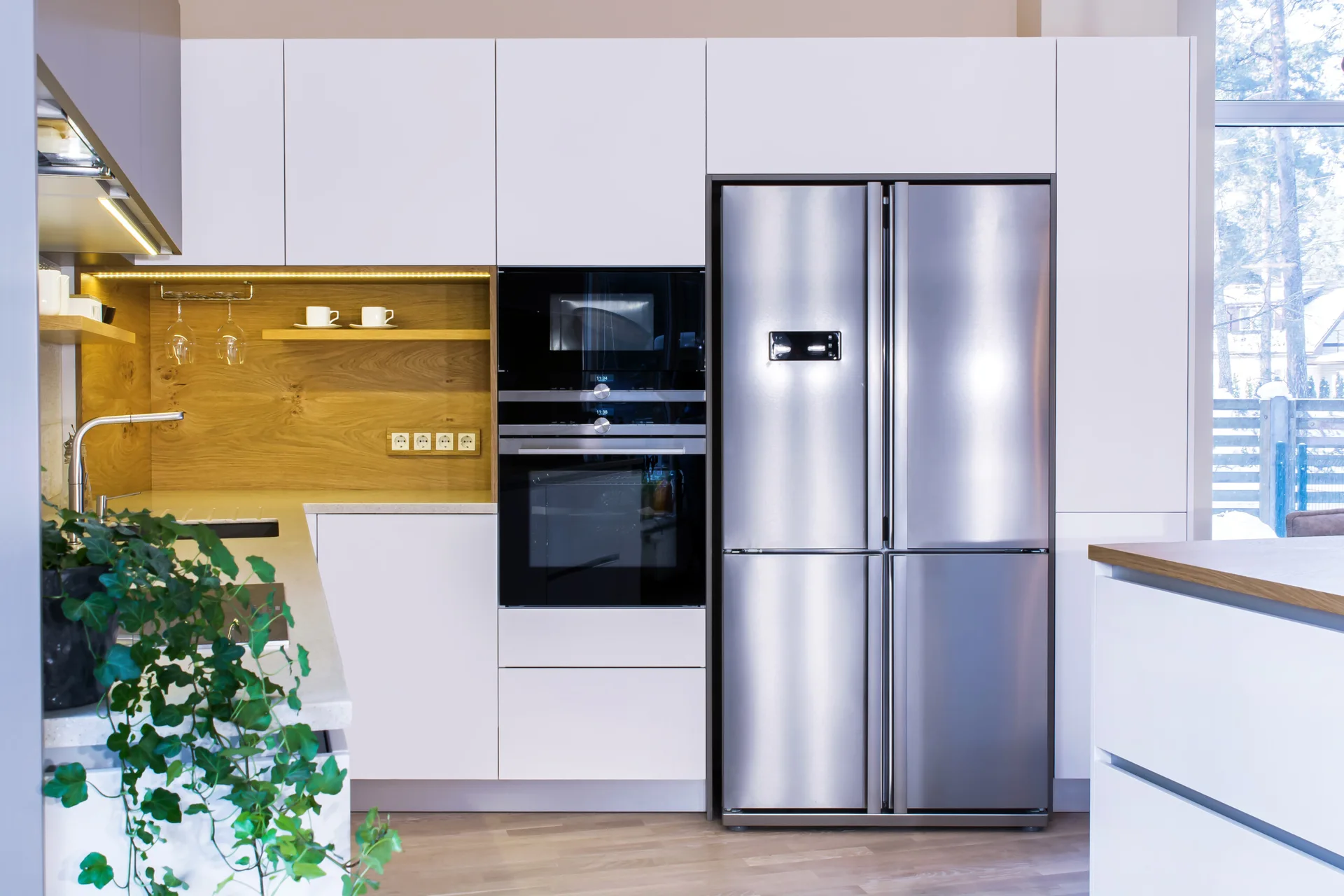 A contemporary kitchen features flat-front white cabinetry surrounding a stainless steel refrigerator, integrated double ovens, and a wood-paneled shelf niche.
