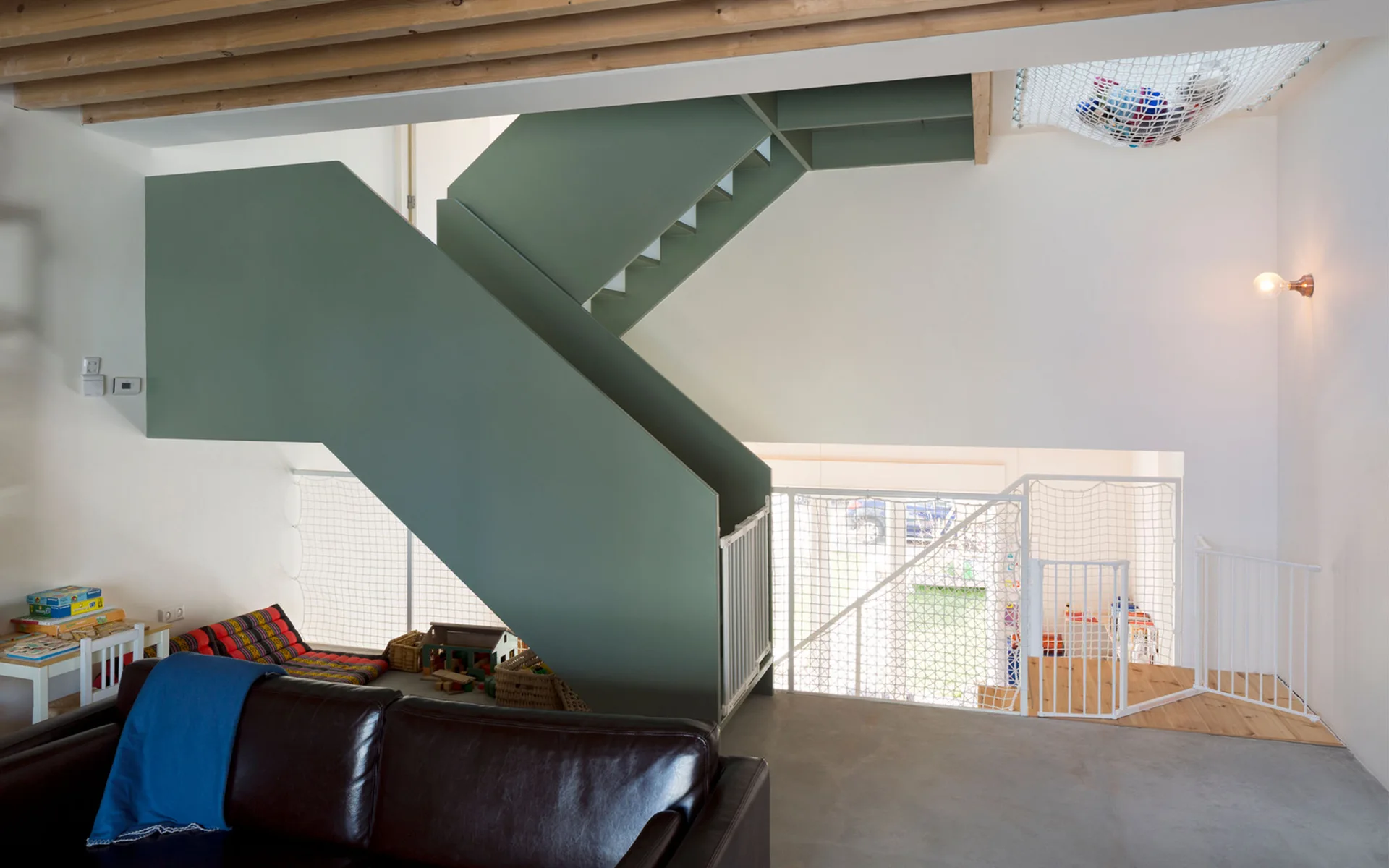 An olive green staircase with white safety netting and baby gates stands in a modern home with wood ceiling beams.
