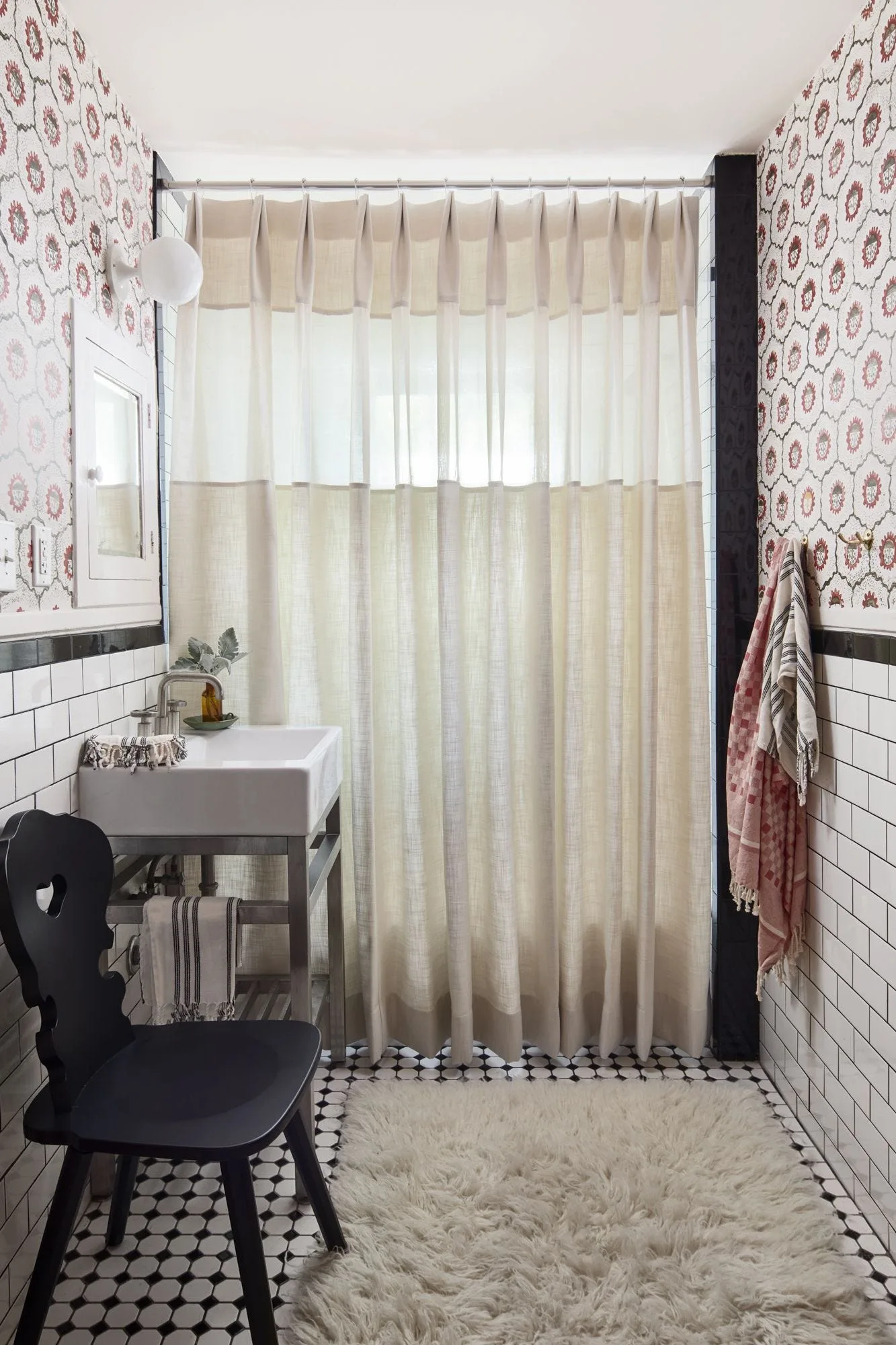 A small bathroom features red patterned wallpaper, white subway tile, a black heart-cutout chair, and a pleated beige shower curtain.