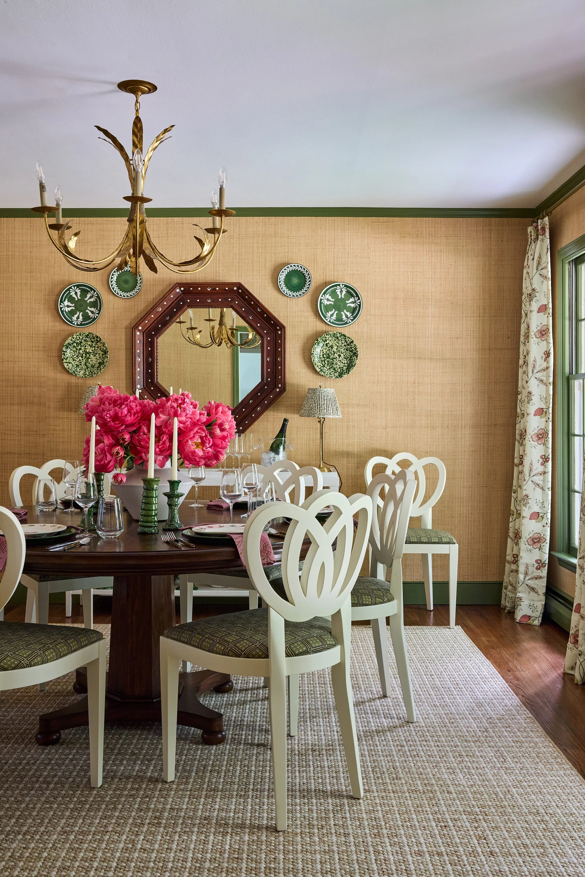 A formal dining room features a round wood table with white chairs, grasscloth wallpaper, green ceiling trim, and a brass chandelier.