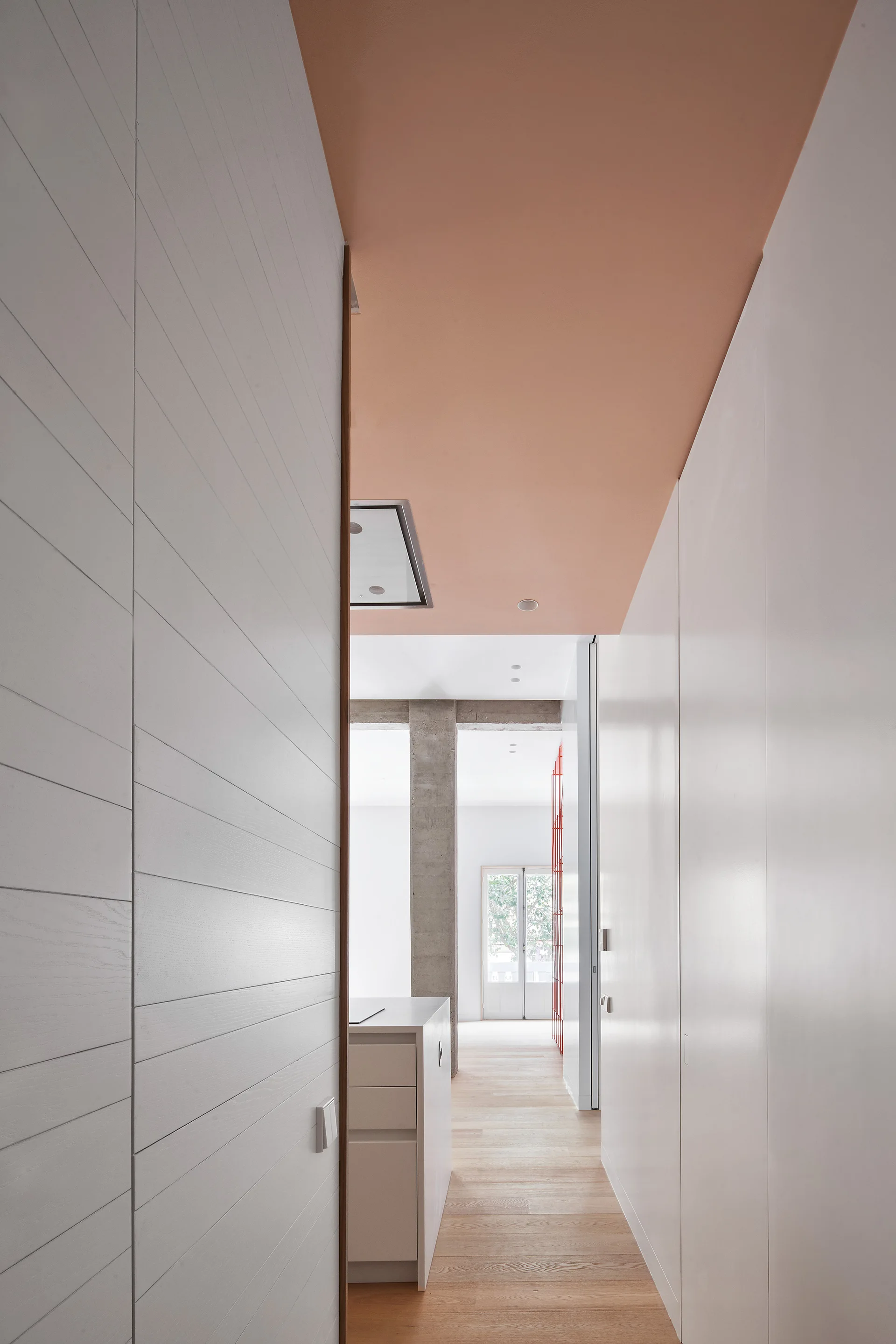 A minimalist white hallway with natural wood floors and a contrasting peach painted ceiling leading to a bright living area.