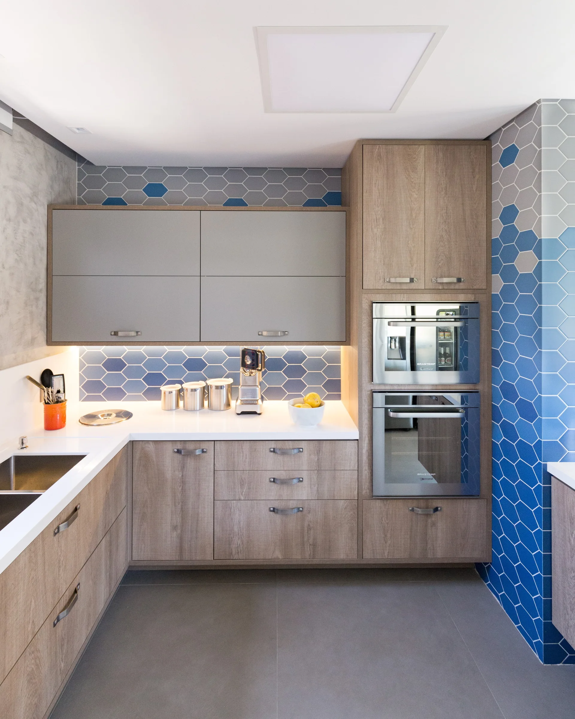 A modern kitchen featuring wood-grain lower cabinets, grey upper cabinets, and a blue and grey hexagonal tile backsplash with under-cabinet lighting.