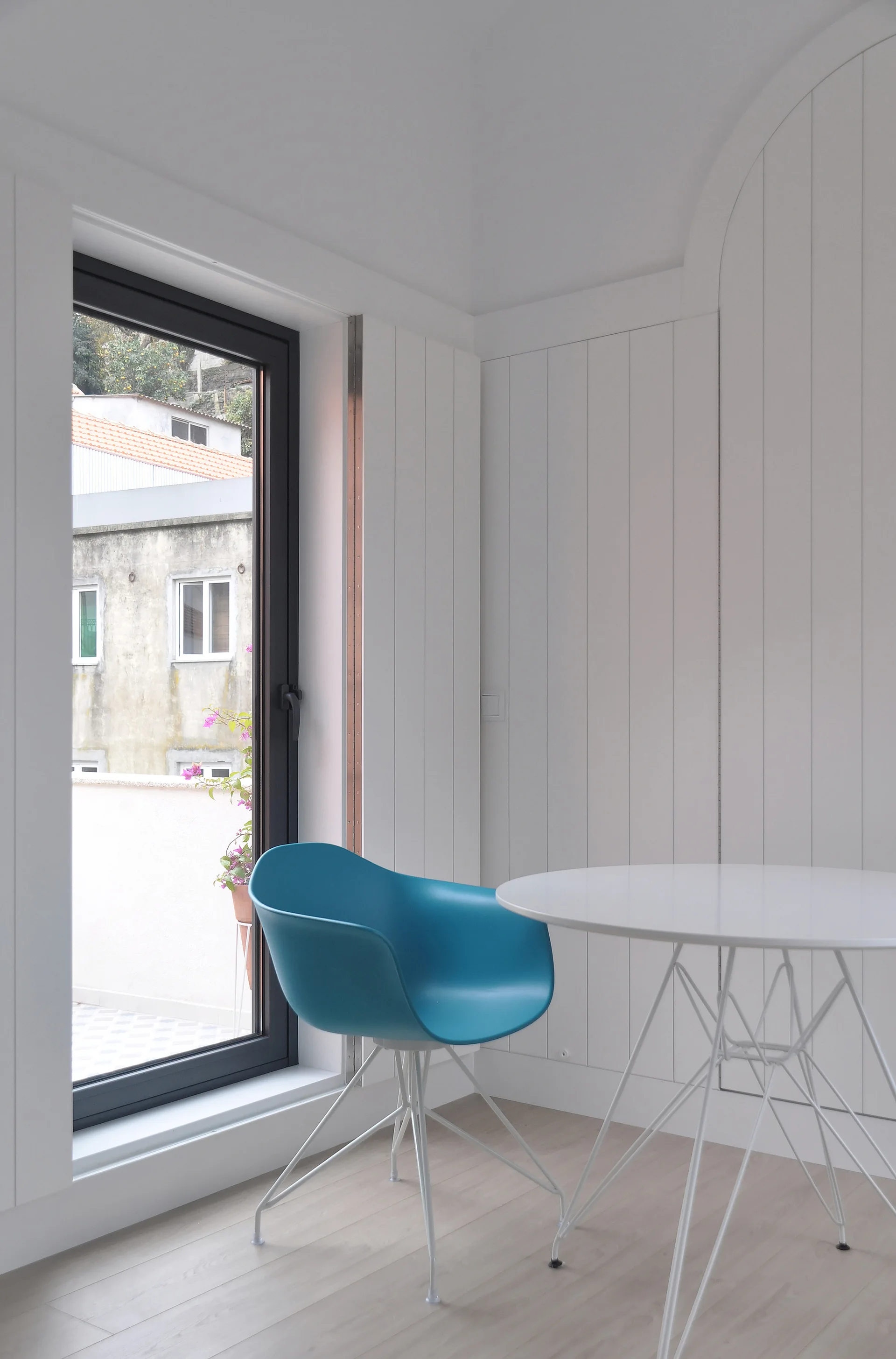 A minimalist corner dining setup with a turquoise plastic armchair and a round white cafe table against white vertical wall paneling.