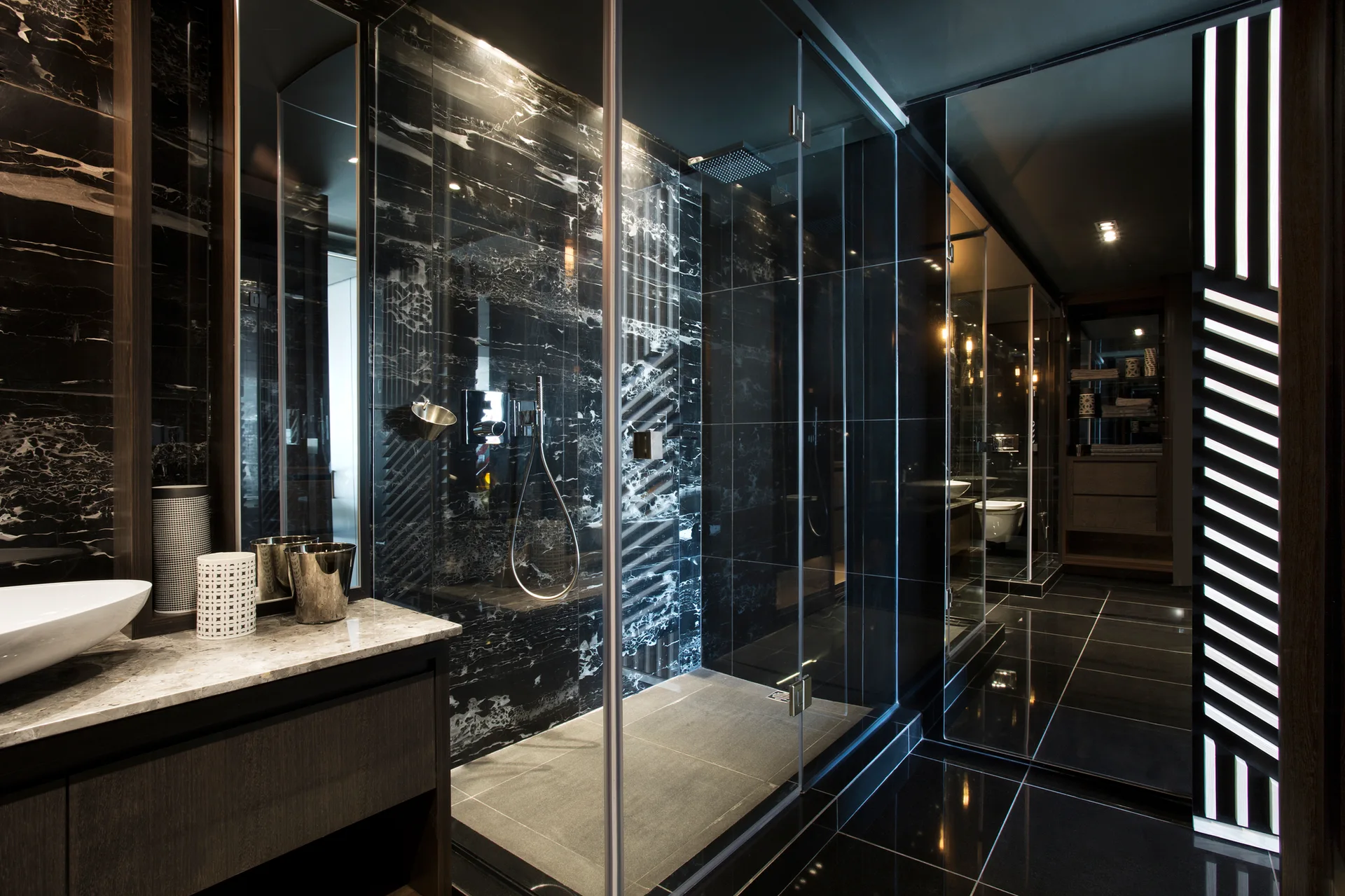 A dark modern bathroom featuring a glass shower with black marble walls, a wood vanity with a white vessel sink, and glossy black floor tiles.