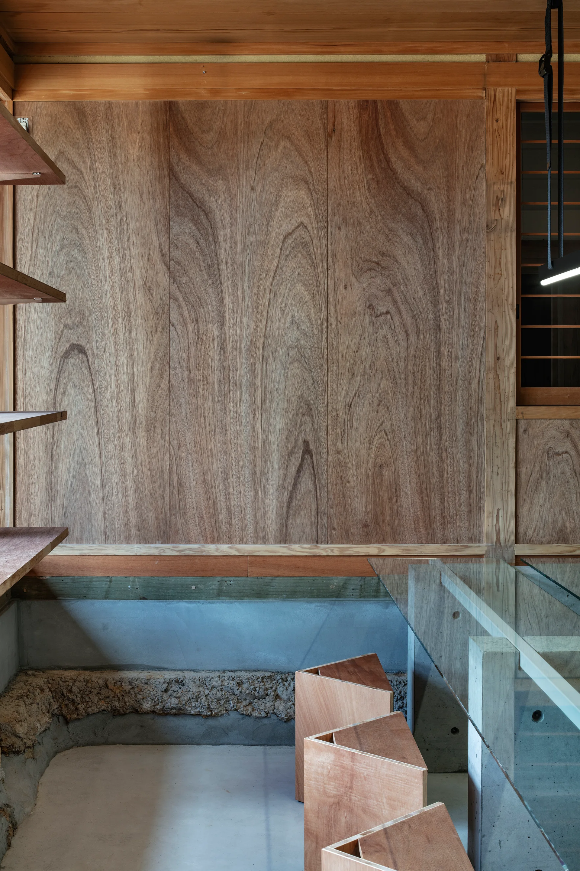 A modern interior space featuring warm wood wall panels, triangular wooden stools, a glass-top table on a concrete base, and exposed foundation elements.