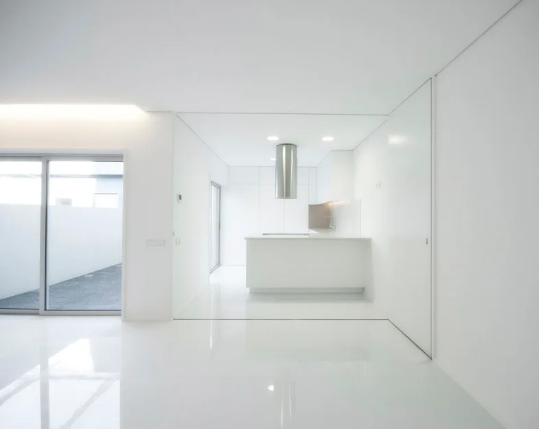 A minimalist all-white kitchen featuring a central island, a cylindrical stainless steel range hood, integrated cabinetry, and high-gloss reflective flooring.
