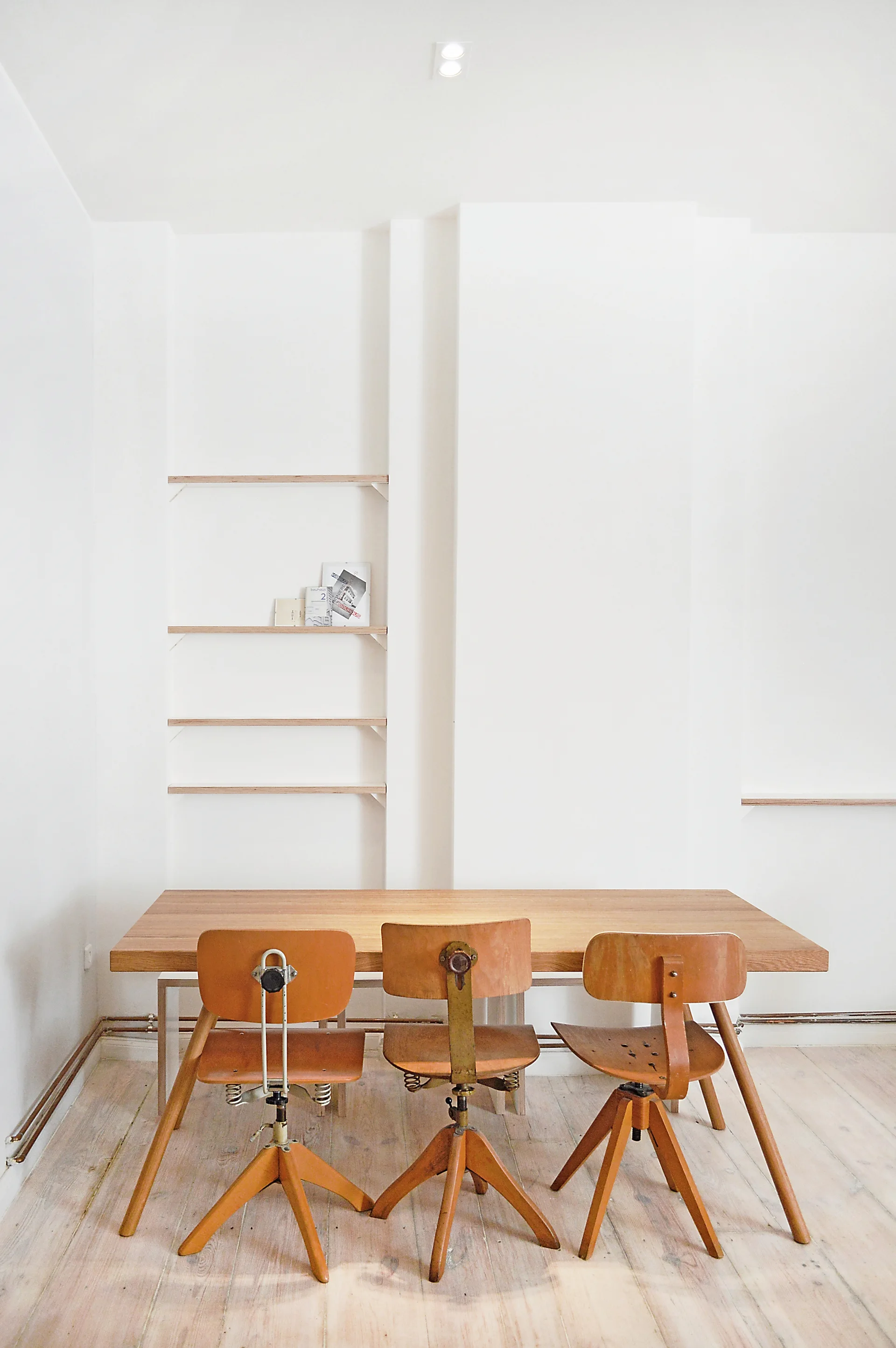 Three vintage wooden office chairs sit at a light wood dining table against a white wall with minimalist recessed shelving.
