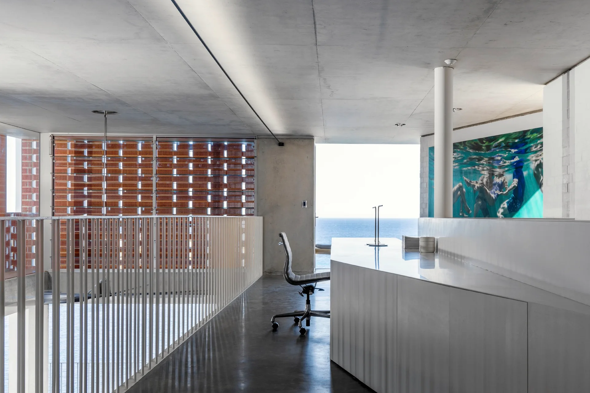 A modernist coastal office with a white geometric desk, grey concrete ceiling and floor, and a terracotta breeze block partition wall.