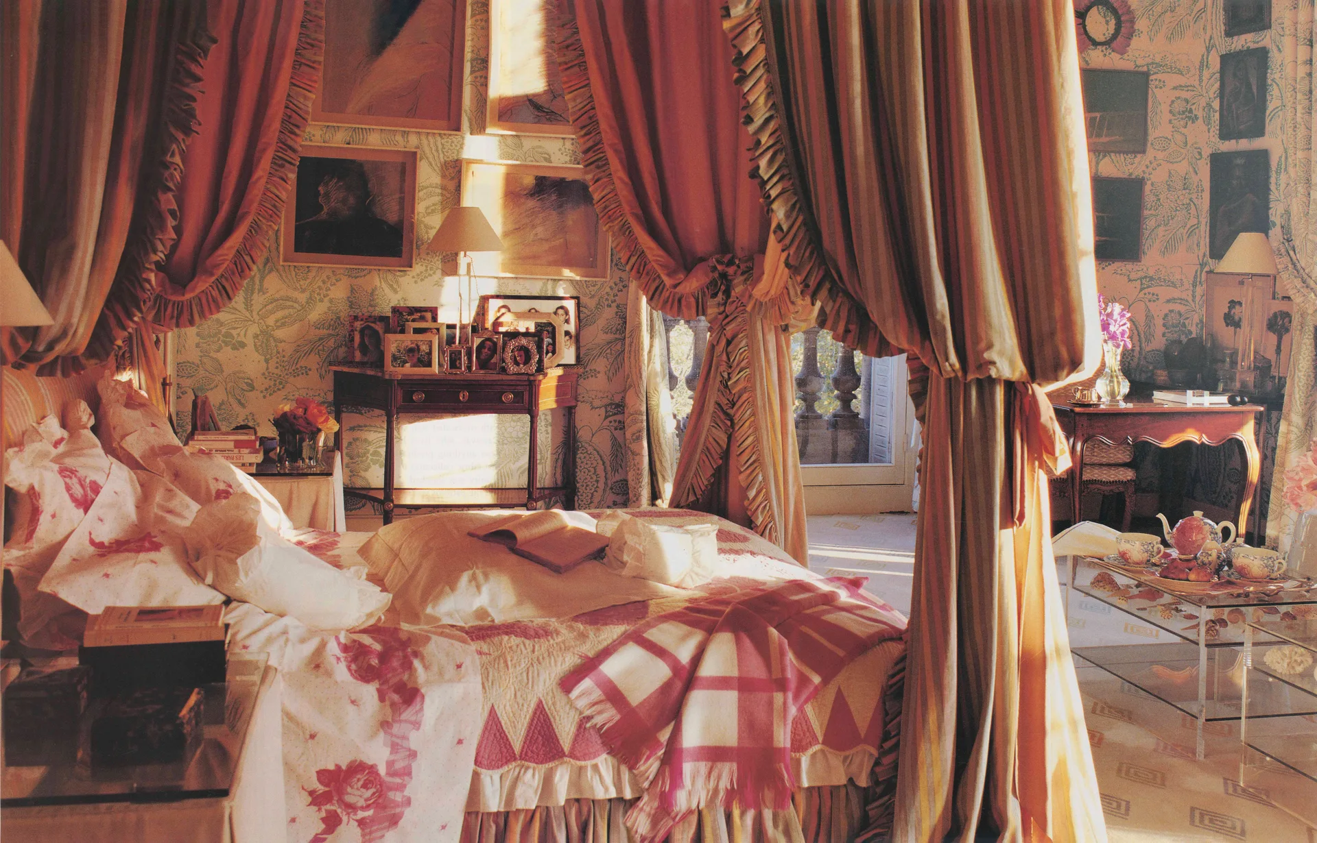 A traditional bedroom features a large canopy bed draped in striped fabric next to green floral wallpaper and a wooden console table with picture frames.