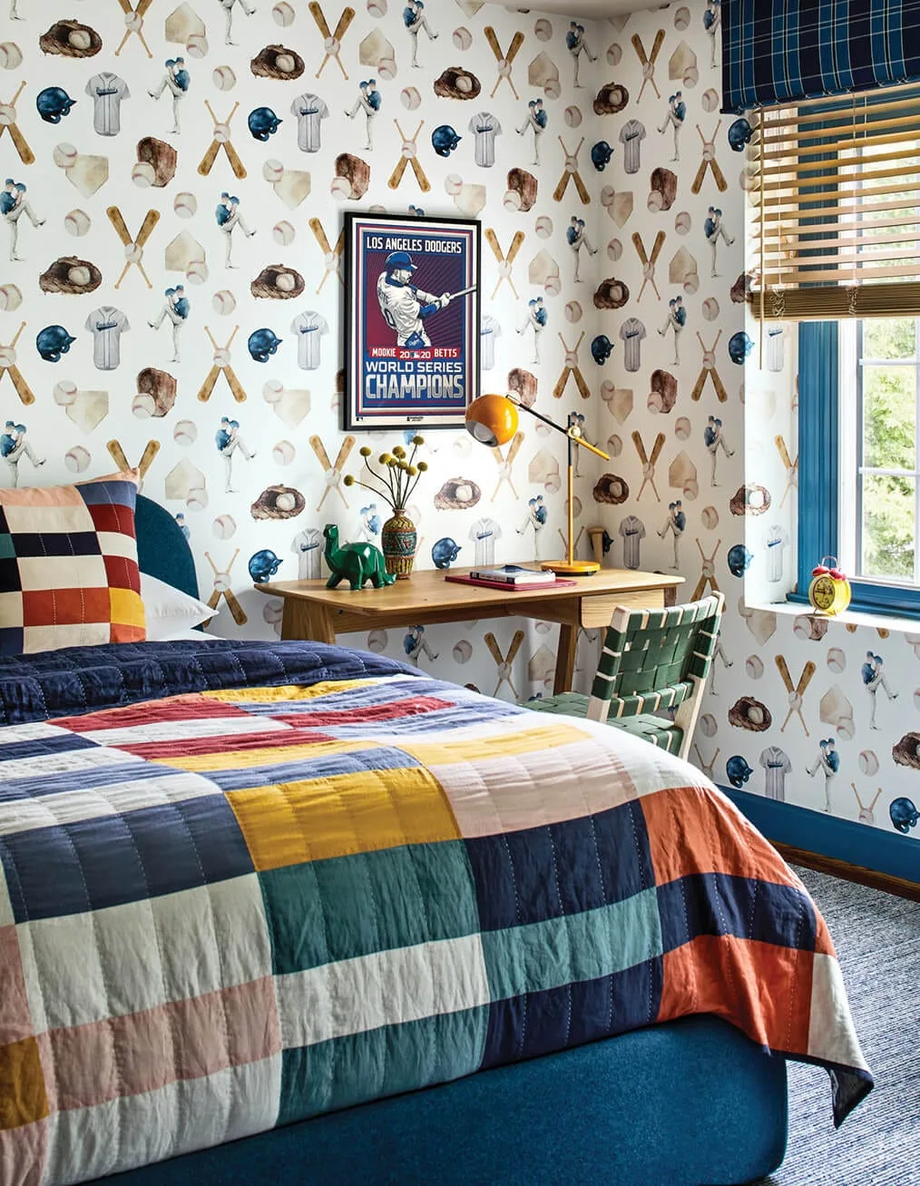 A children's bedroom features baseball-themed wallpaper, a wooden desk with a yellow lamp, and a colorful patchwork quilt on the bed.