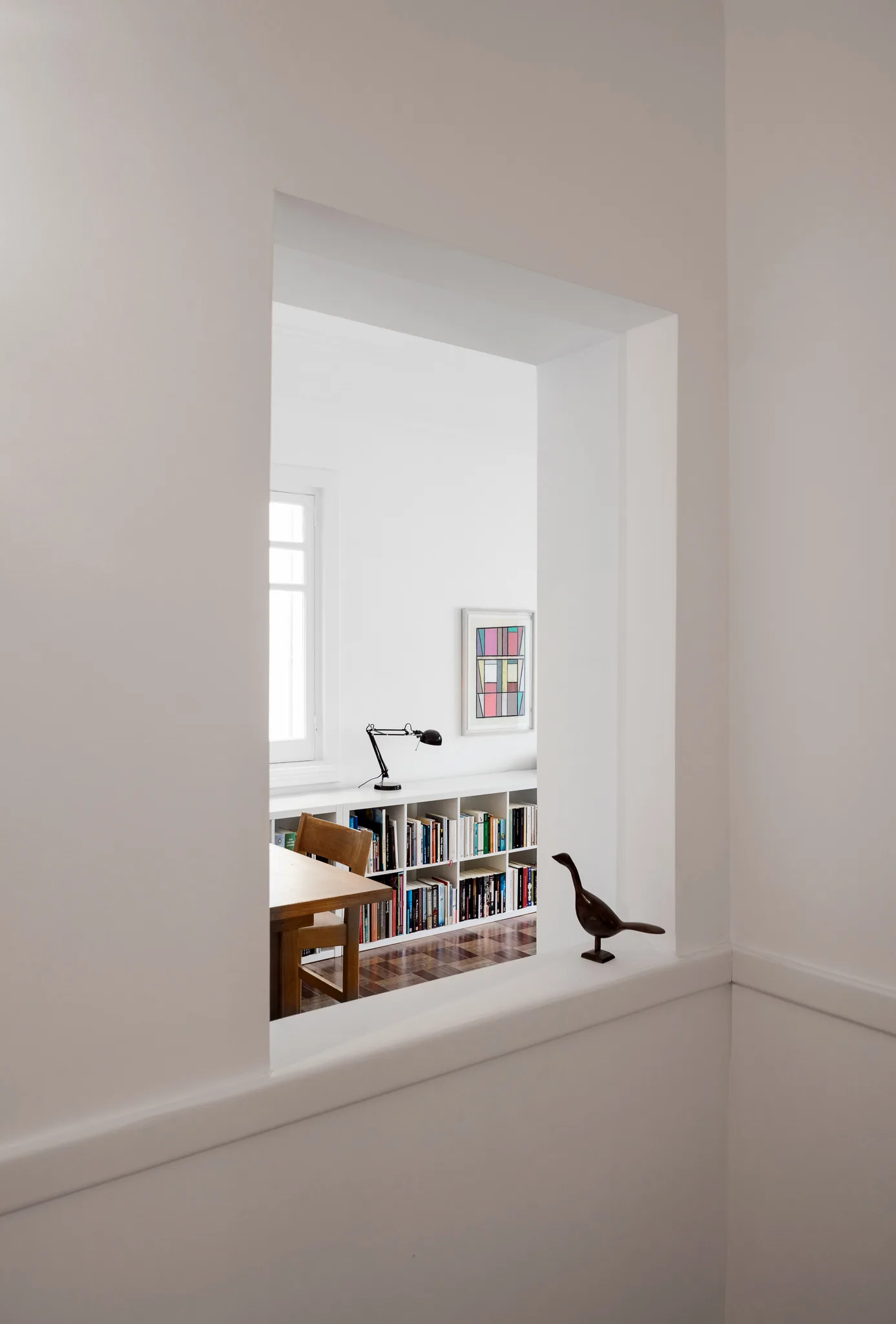 A view through a white rectangular wall opening reveals a minimalist home office with a wooden desk, low bookshelf, and black task lamp.