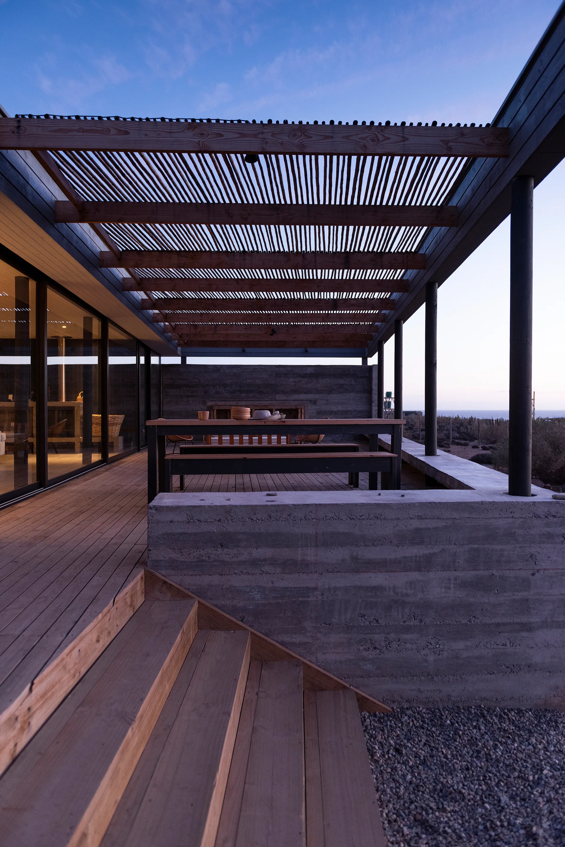 A wooden deck patio featuring an outdoor dining table under a slatted pergola roof alongside a board-formed concrete retaining wall.