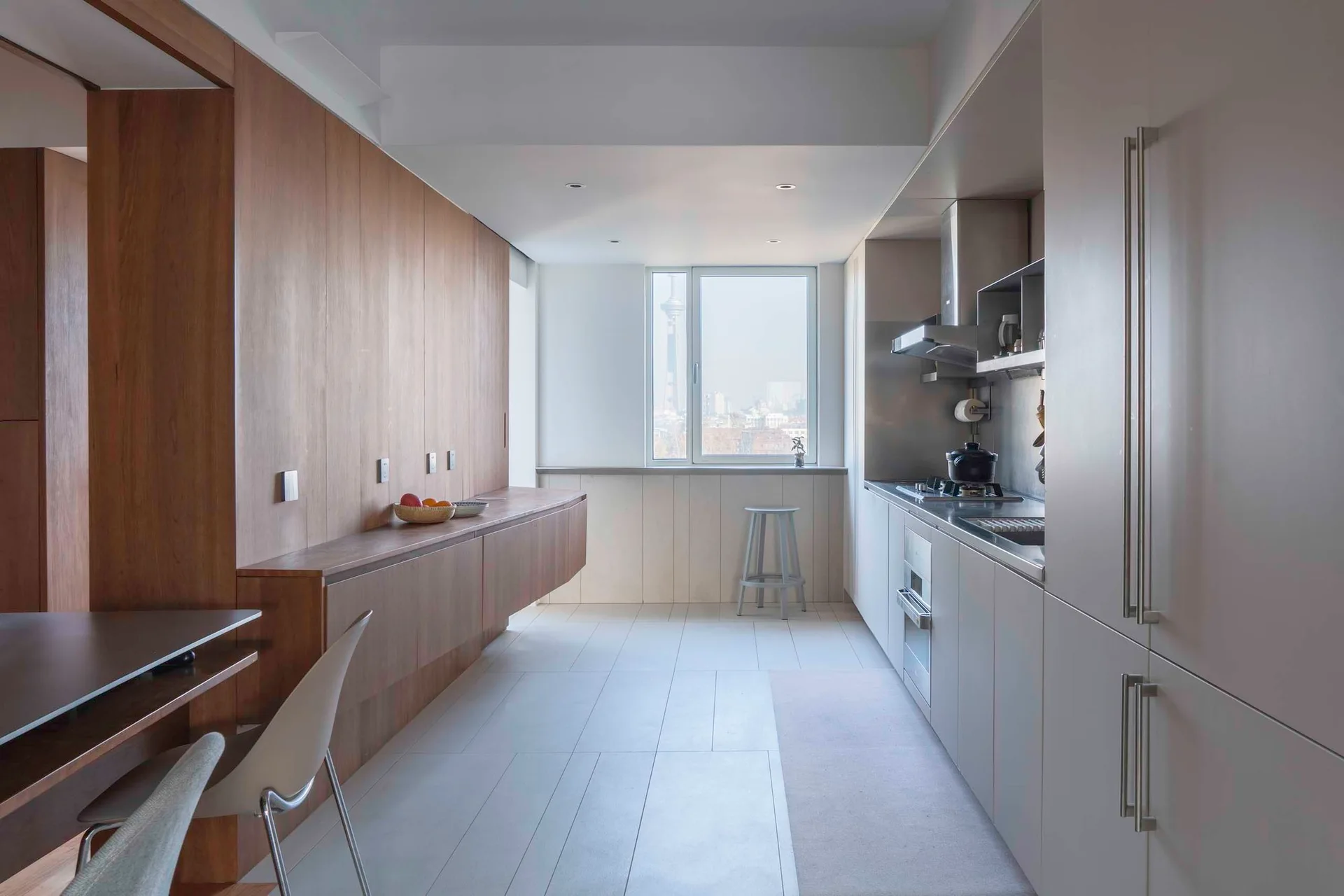 A minimalist galley kitchen features warm wood wall paneling on one side and sleek grey cabinetry on the other, leading to a window with a city view.