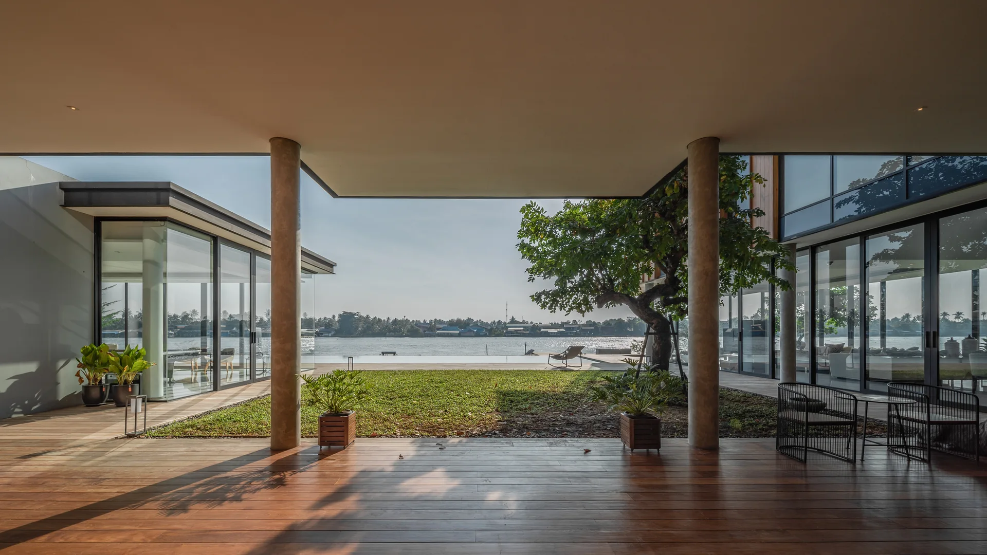 A covered wooden patio with round concrete columns overlooks a grassy lawn and a wide river under a clear sky.