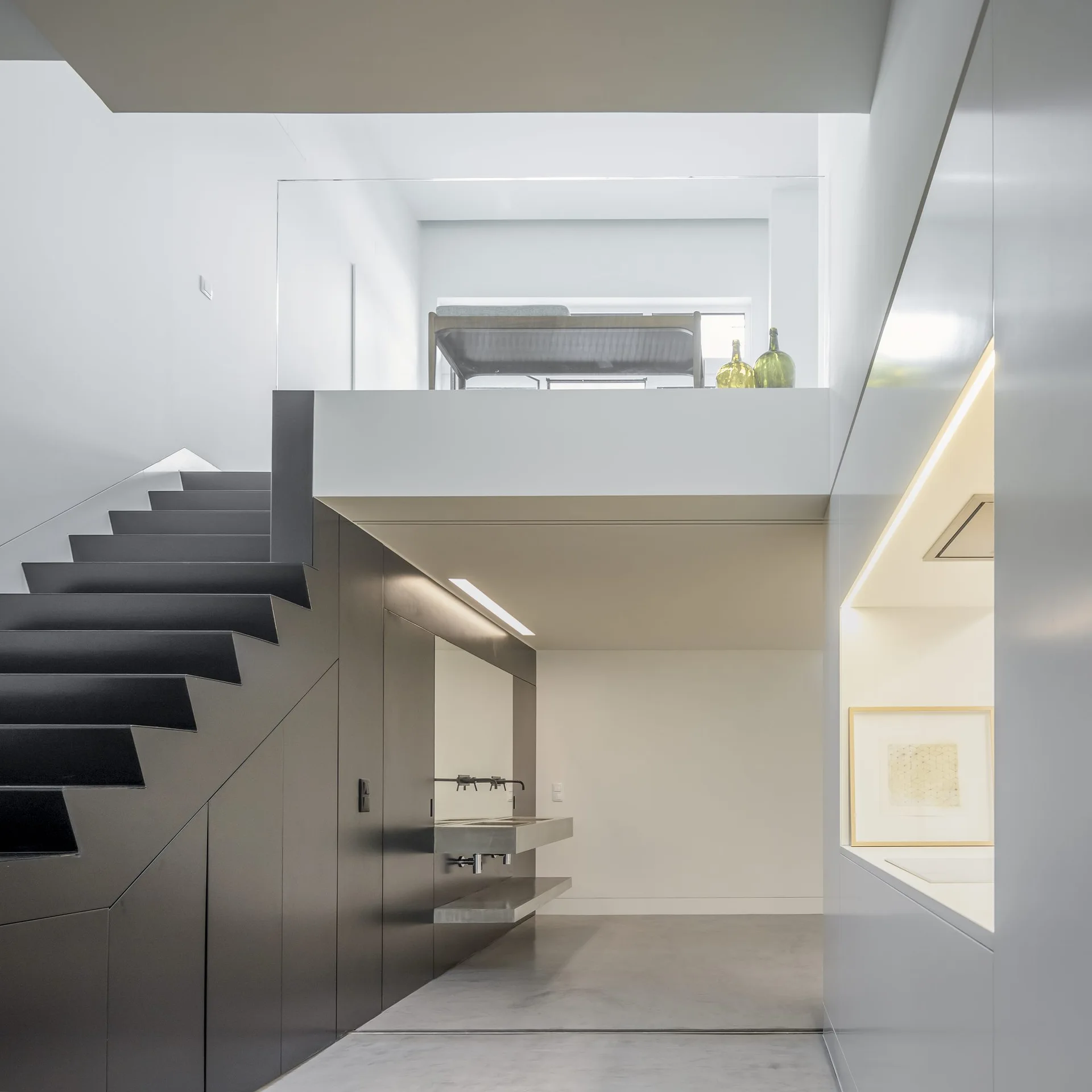 A minimalist multi-level loft features a dark angular staircase, a concrete floating vanity, and a mezzanine level with a glass railing.