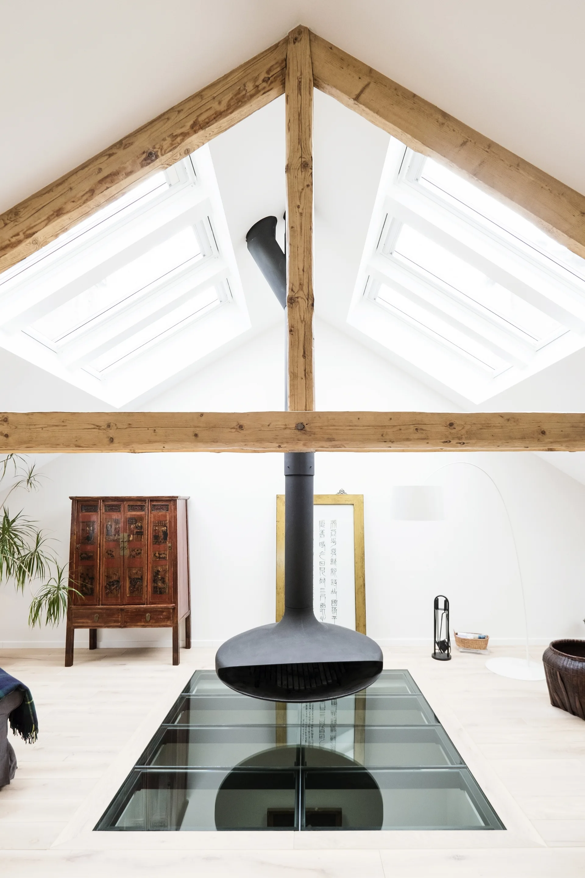 A bright attic living room featuring a black suspended fireplace above a glass floor panel and beneath a vaulted ceiling with wooden beams.