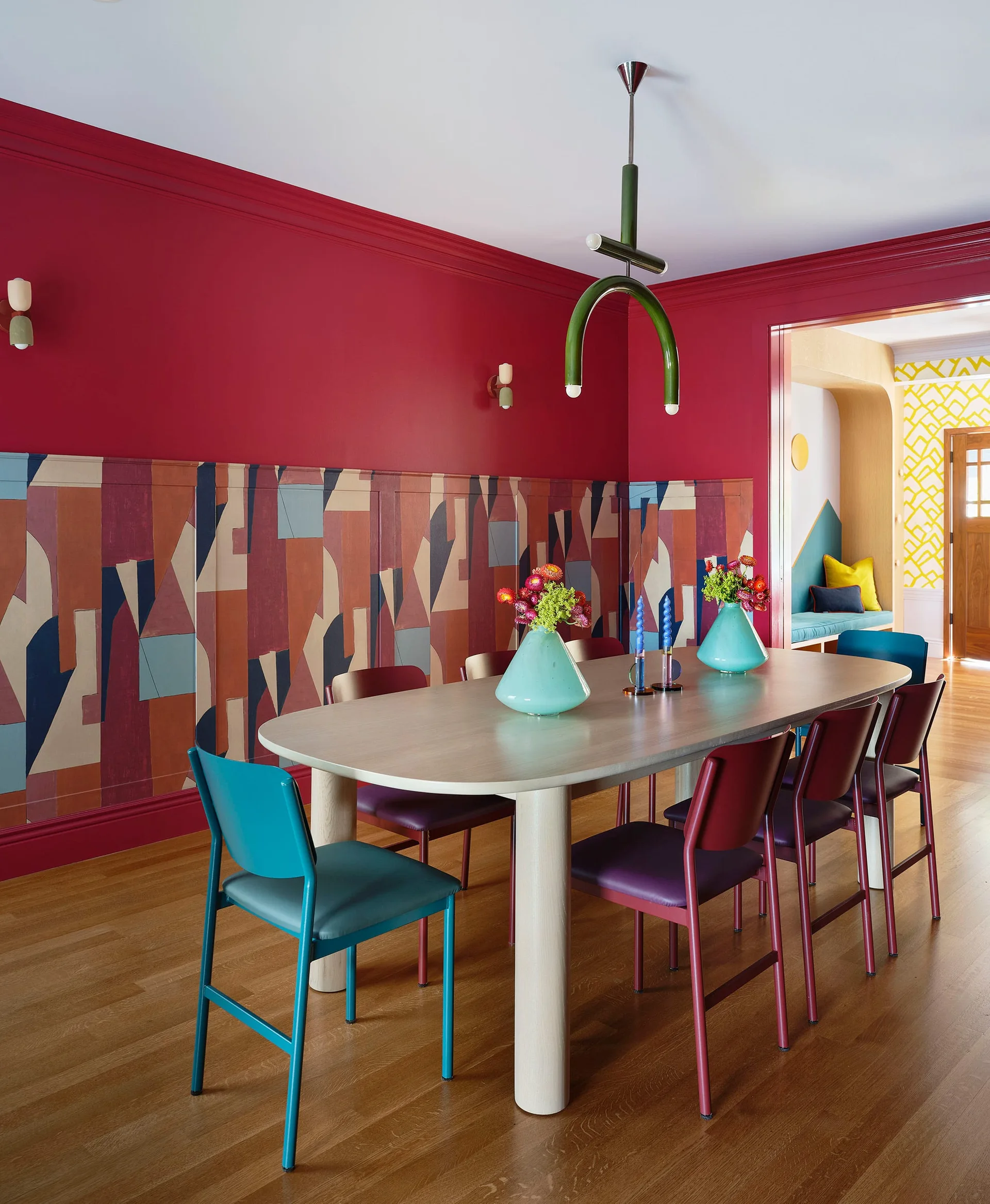 A modern dining room featuring a light wood table and colorful chairs against a deep red wall with geometric patterned wainscoting.