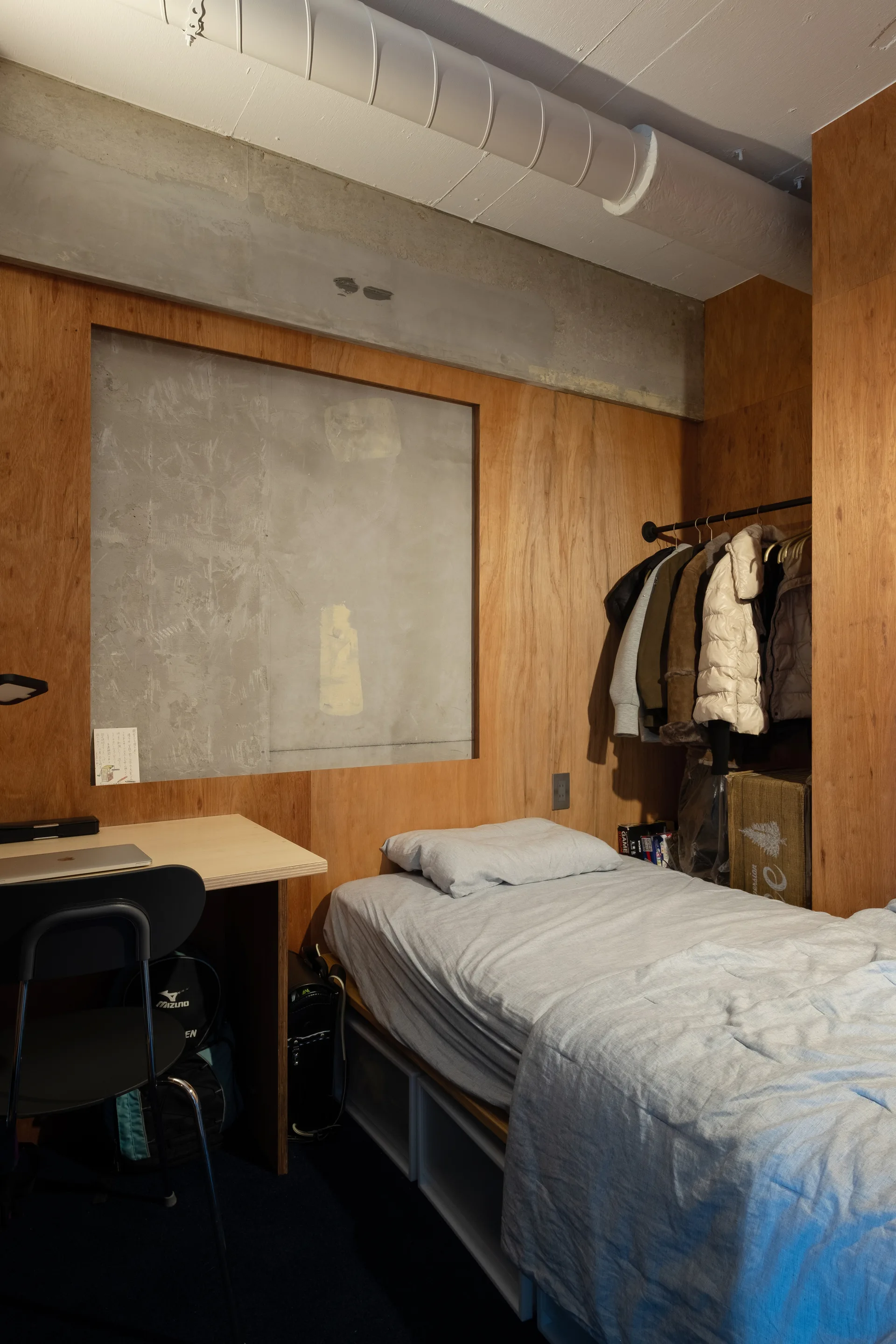 A small minimalist bedroom featuring warm plywood paneled walls, an exposed concrete ceiling section, a single bed, and a simple study desk.