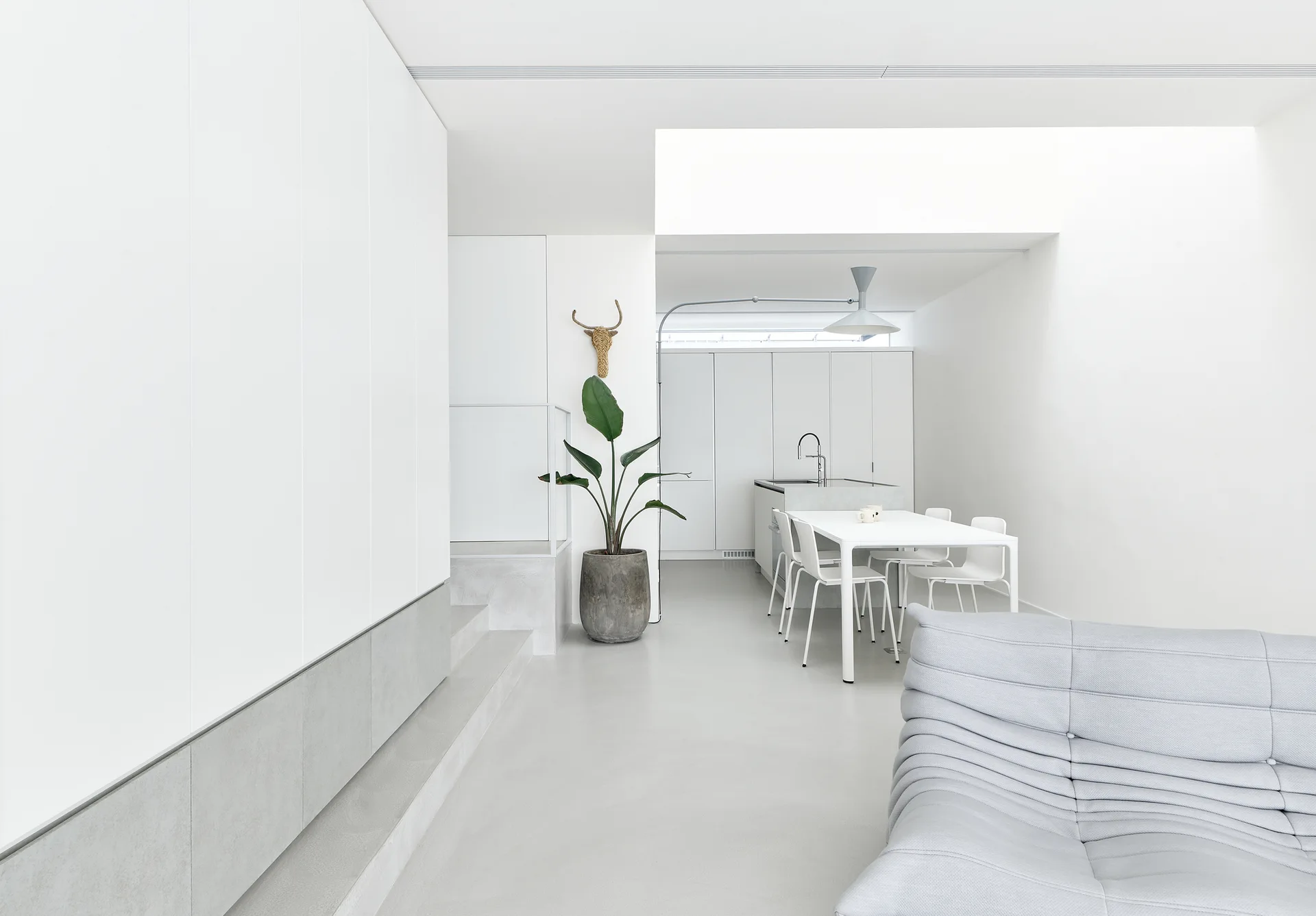 A bright minimalist open-concept space featuring a white dining table, light grey flooring, seamless white kitchen cabinets, and a grey tufted sofa.