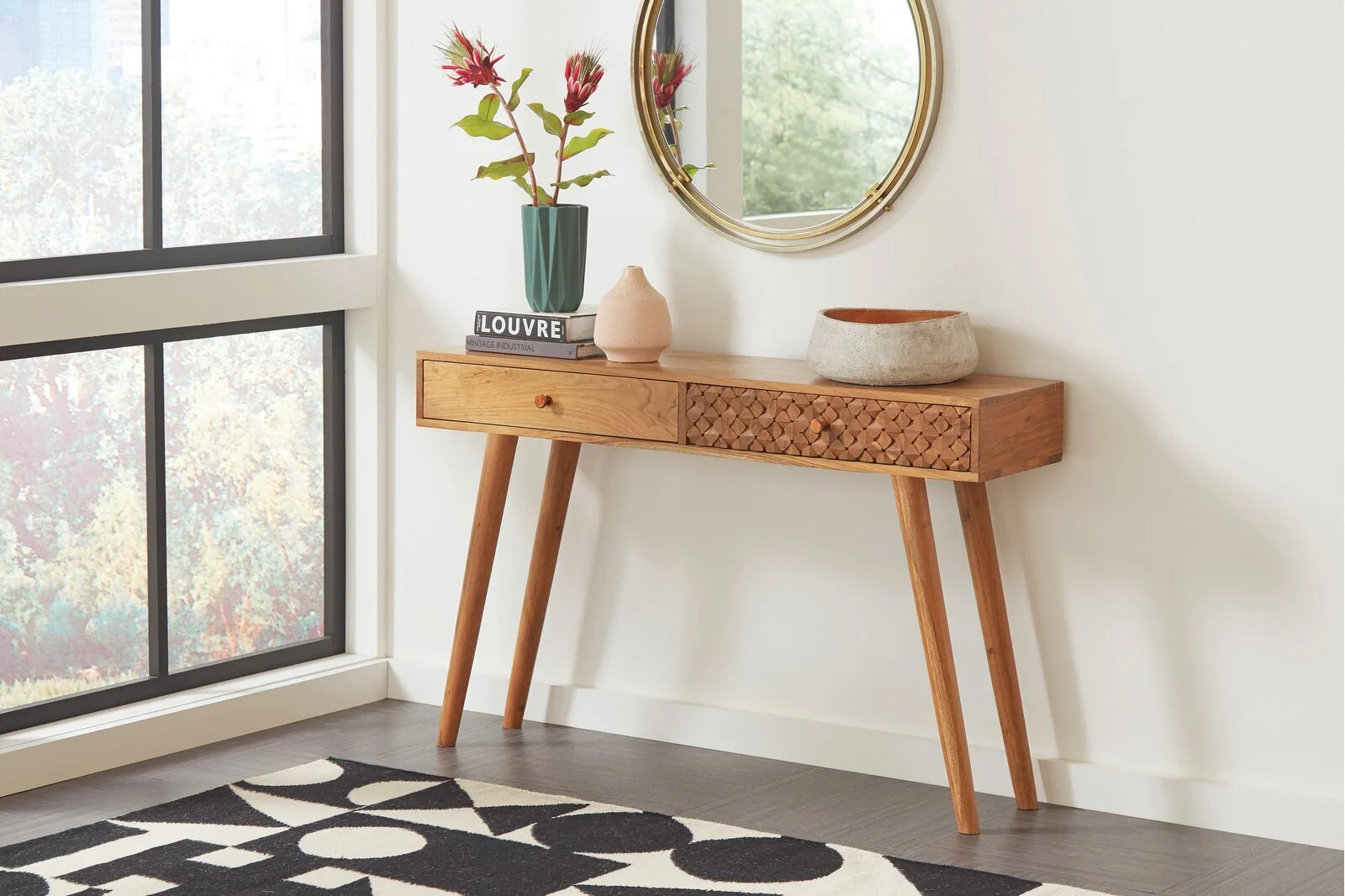 A mid-century wooden console table with a textured drawer front features a round gold mirror above it and decorative vases on top.