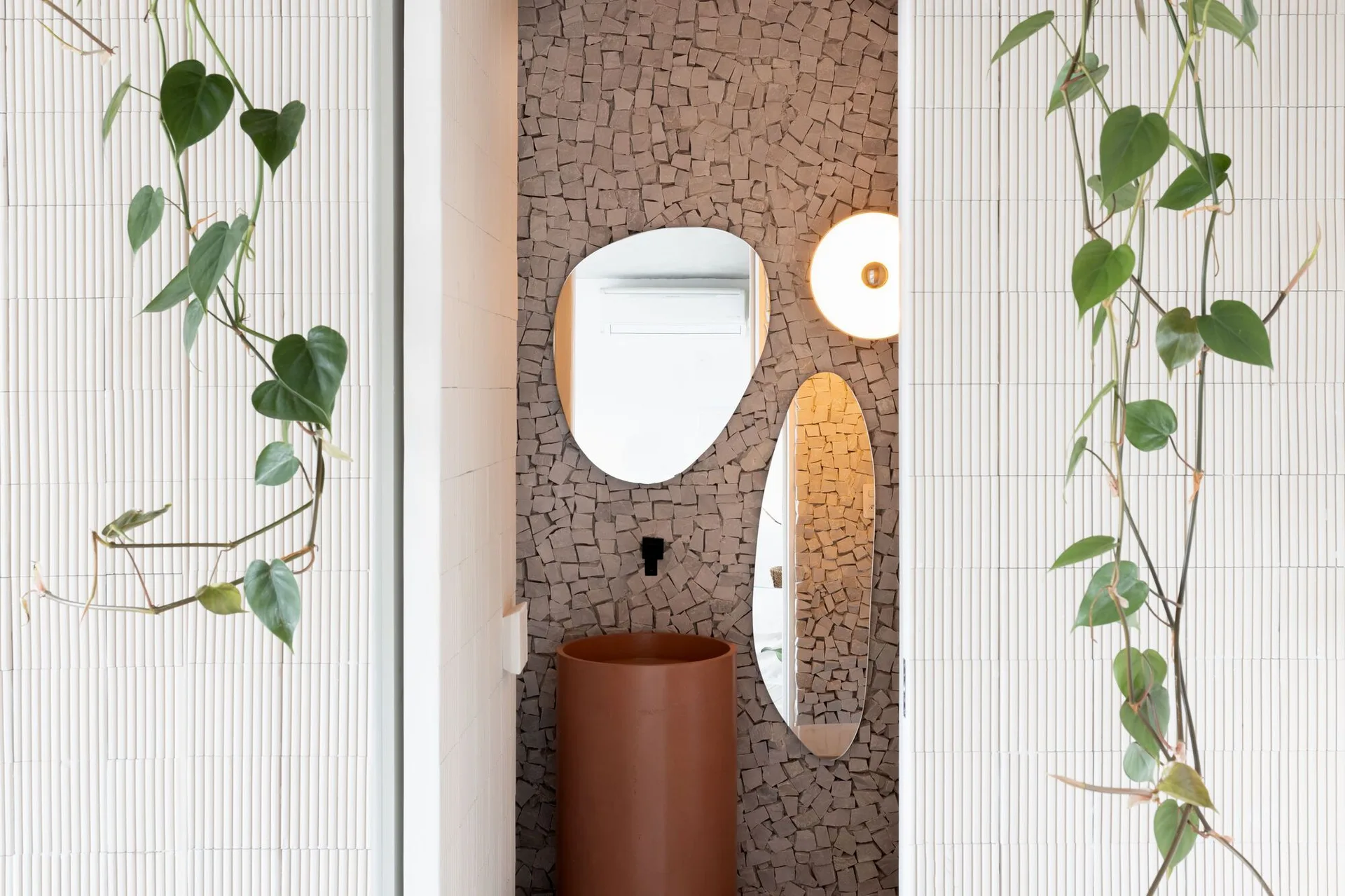 A view into a powder room featuring a cylindrical terracotta pedestal sink, an irregular stone wall, asymmetrical mirrors, and hanging green vines.