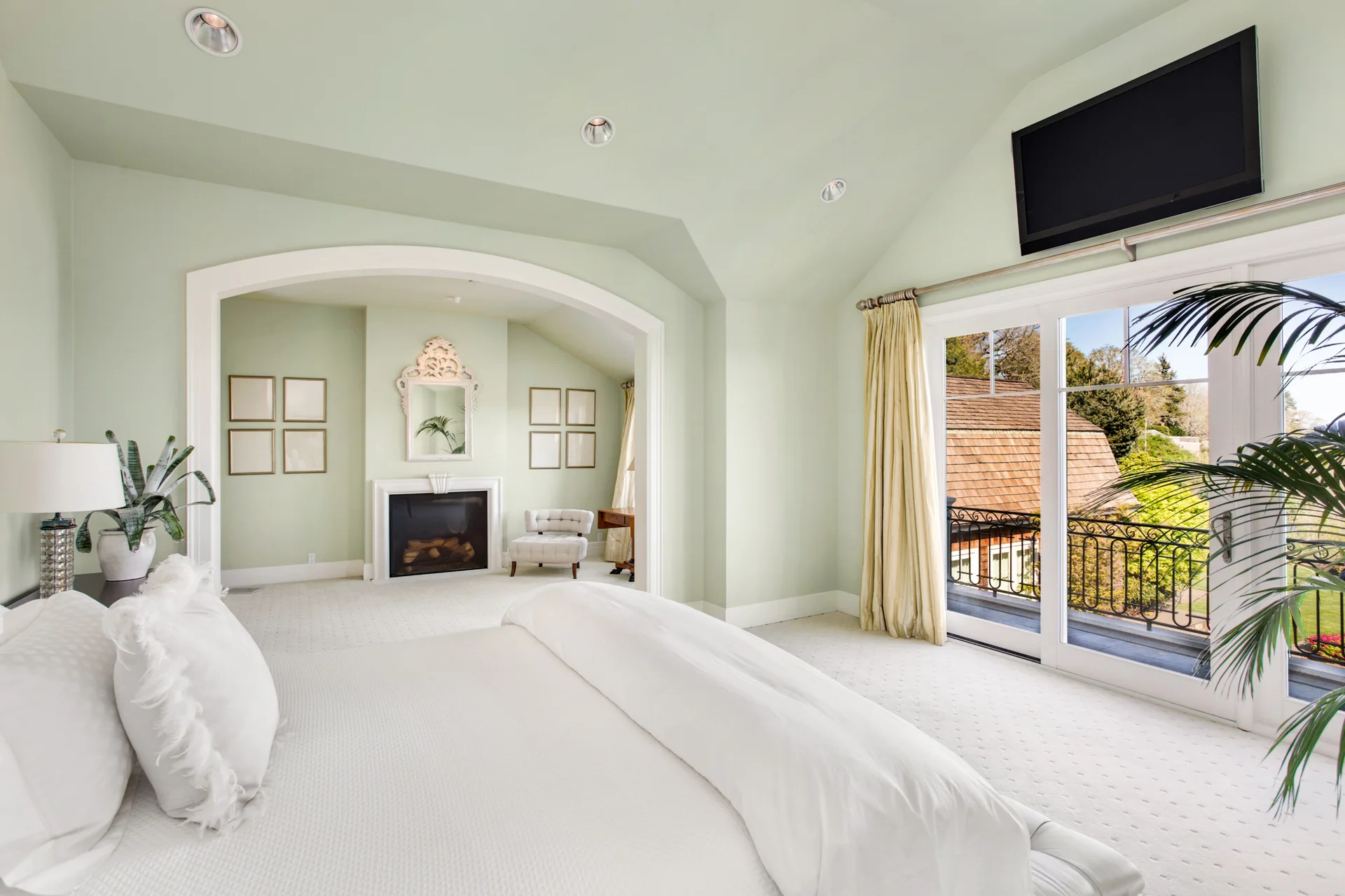 A spacious bedroom with mint green walls and a vaulted ceiling features a white bed, glass balcony doors, and an arched entryway to a fireplace nook.