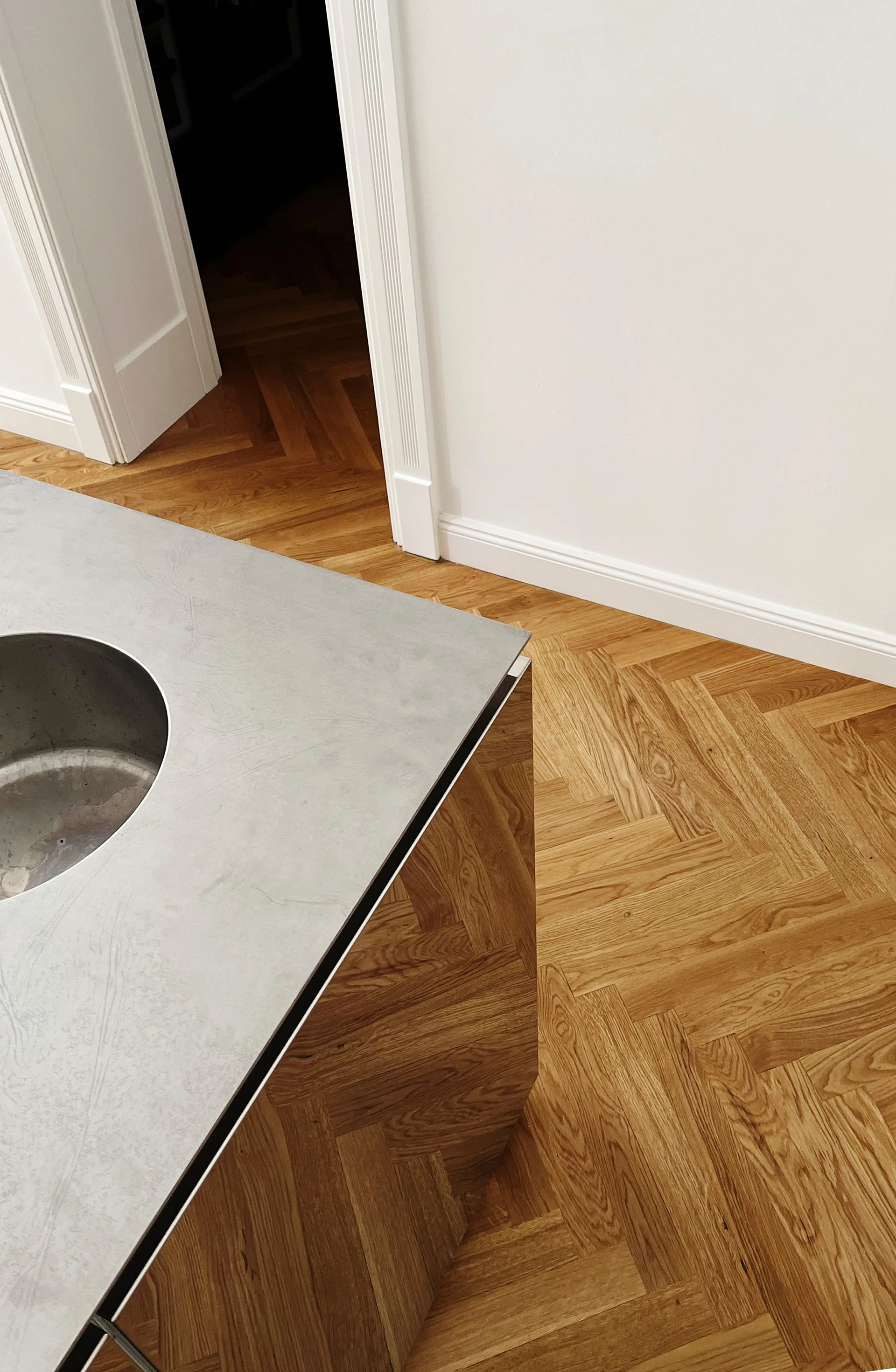 Close-up of a kitchen island with a grey stone countertop and wood paneling on an oak herringbone hardwood floor.