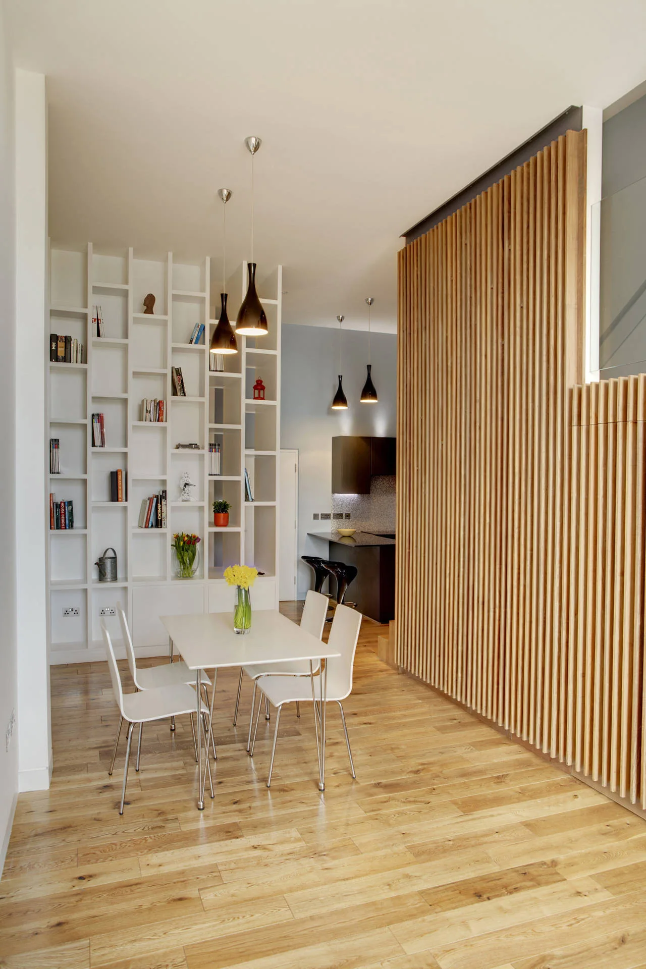 A dining area features a white table and chairs next to a large white bookshelf and a vertical wood slat partition screen on wood flooring.