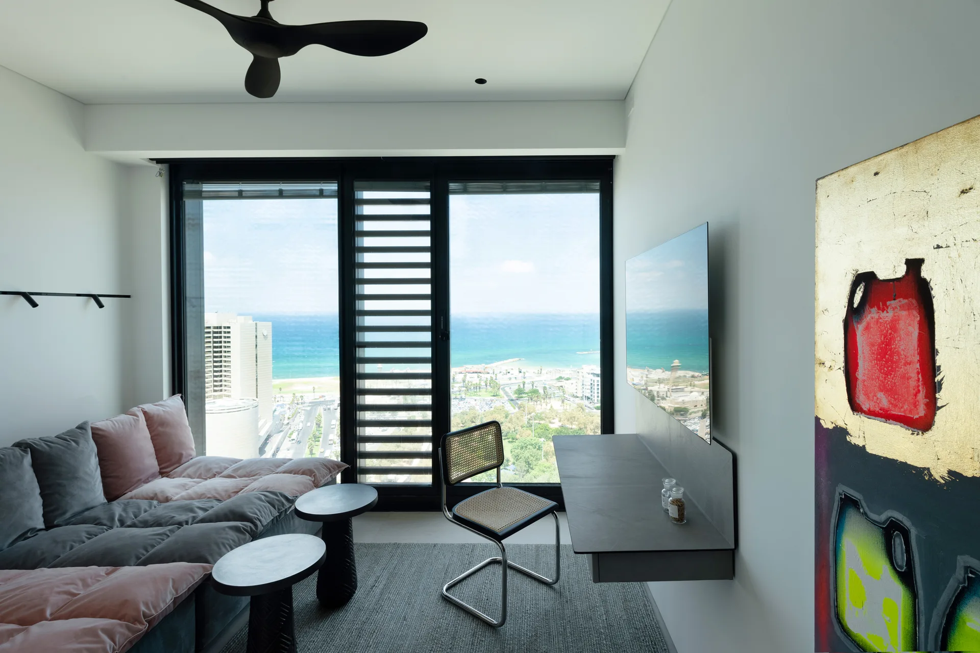 A minimalist room features a grey modular sofa, a wall-mounted floating desk with a cane chair, and a large window looking out at the ocean.