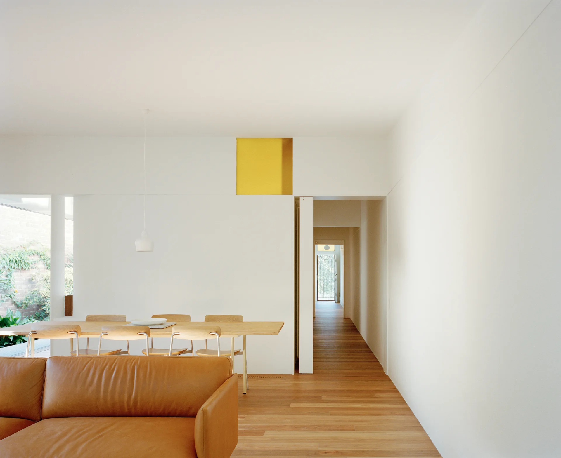 A minimalist living room features a tan leather sofa, a light wood dining set, and a high yellow square panel built into a white partition wall.