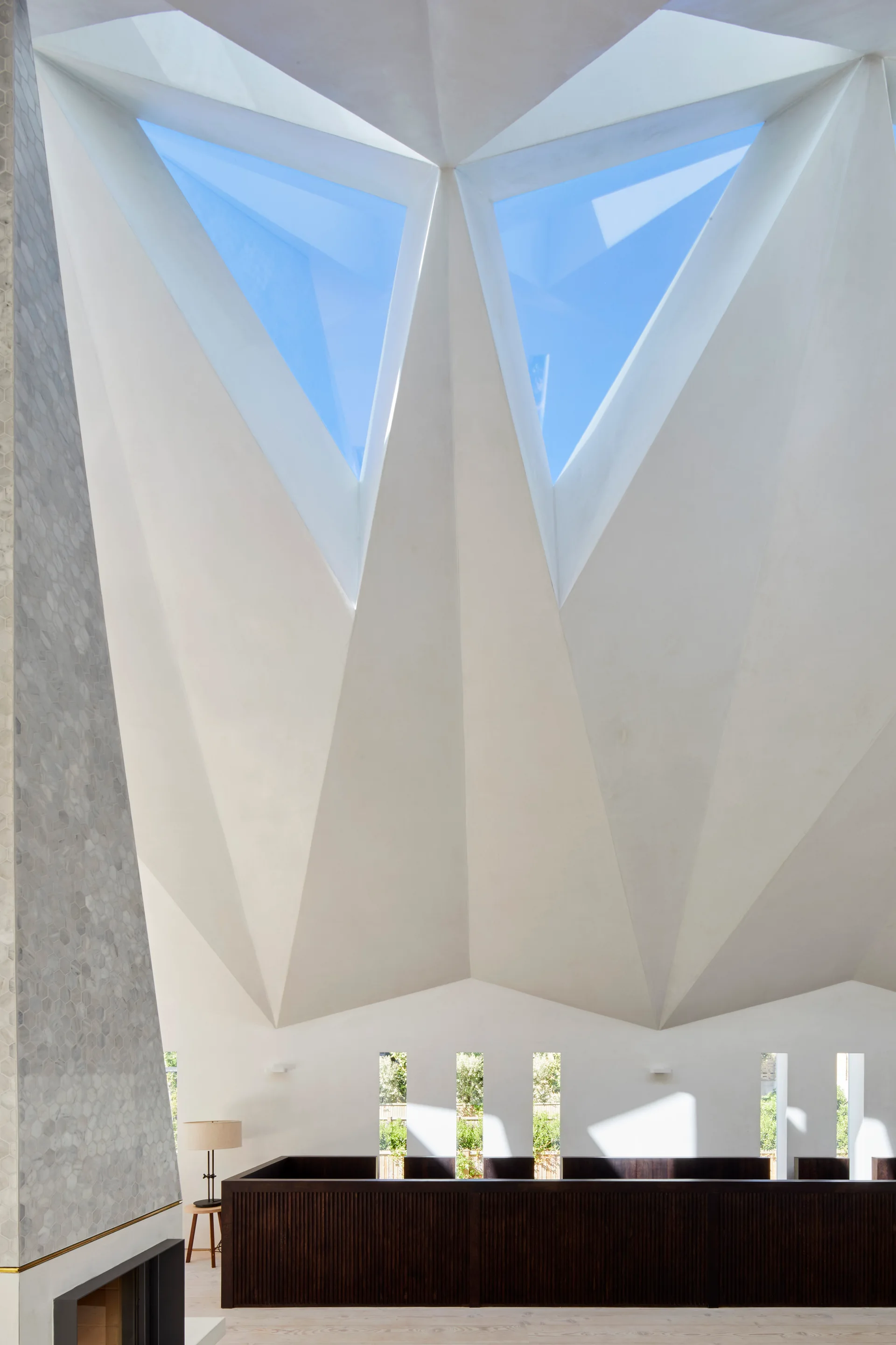 A white interior room showcases a high geometric vaulted ceiling with triangular skylights above a dark wood slatted partition.