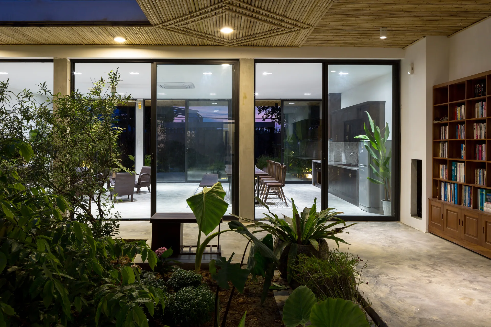 An indoor garden courtyard with green plants is separated by floor-to-ceiling glass sliding doors from a dining area and kitchen.