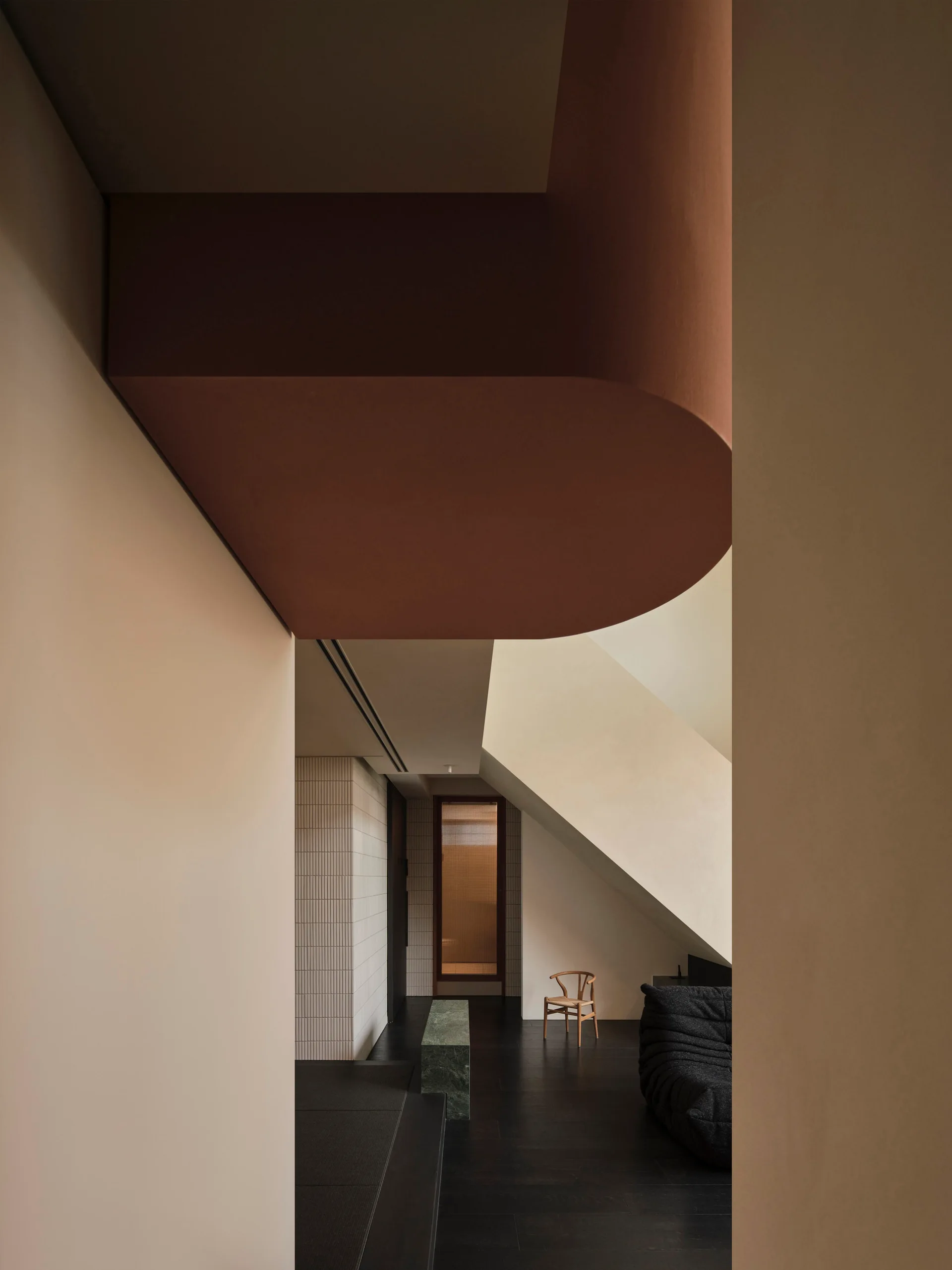 A minimalist hallway features a curved terracotta ceiling element, dark wood floors, a green stone bench, and a distant wooden chair.