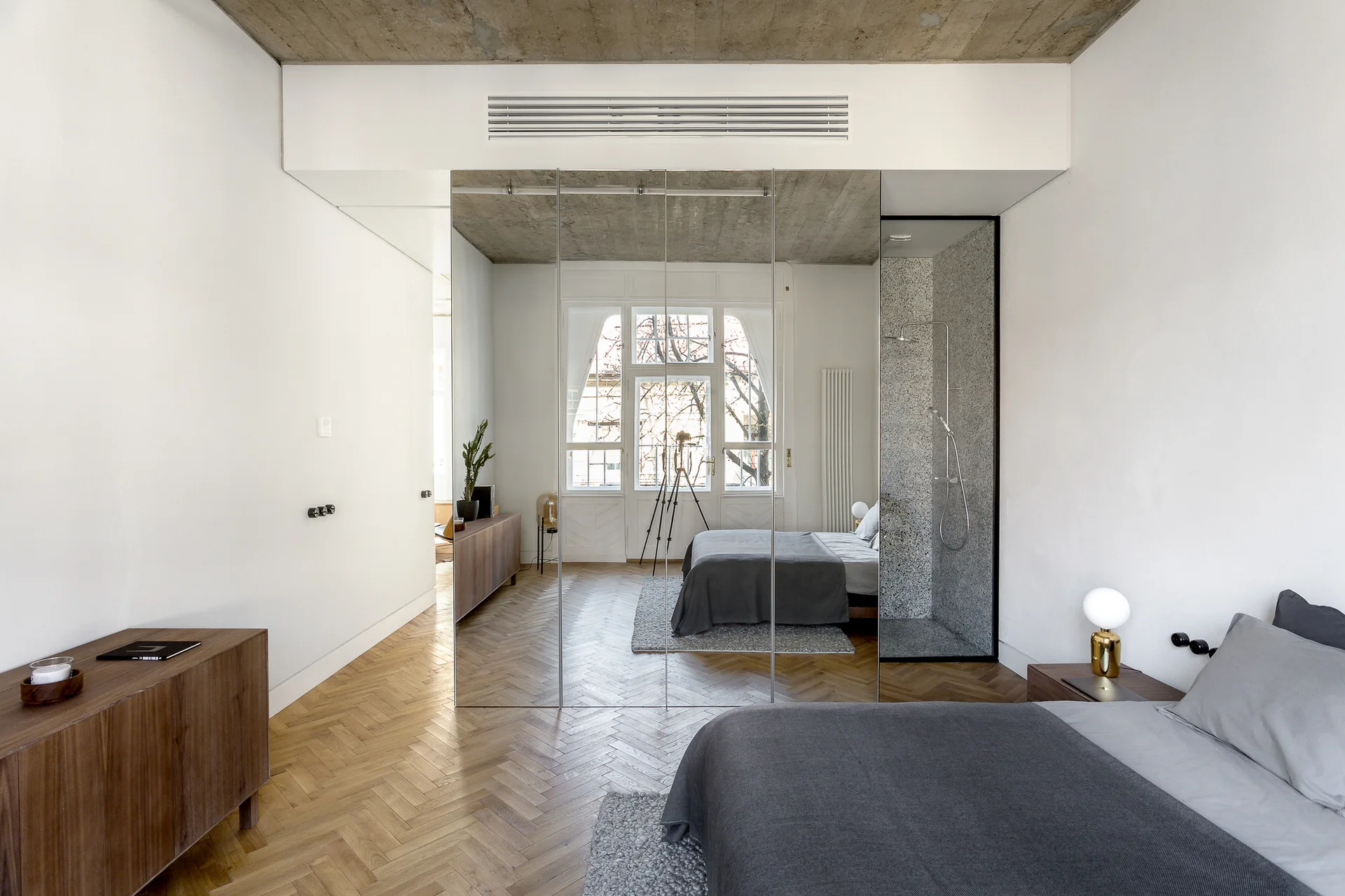 A minimalist bedroom features herringbone wood floors, a concrete ceiling, and a large floor-to-ceiling mirrored partition wall next to a bed.