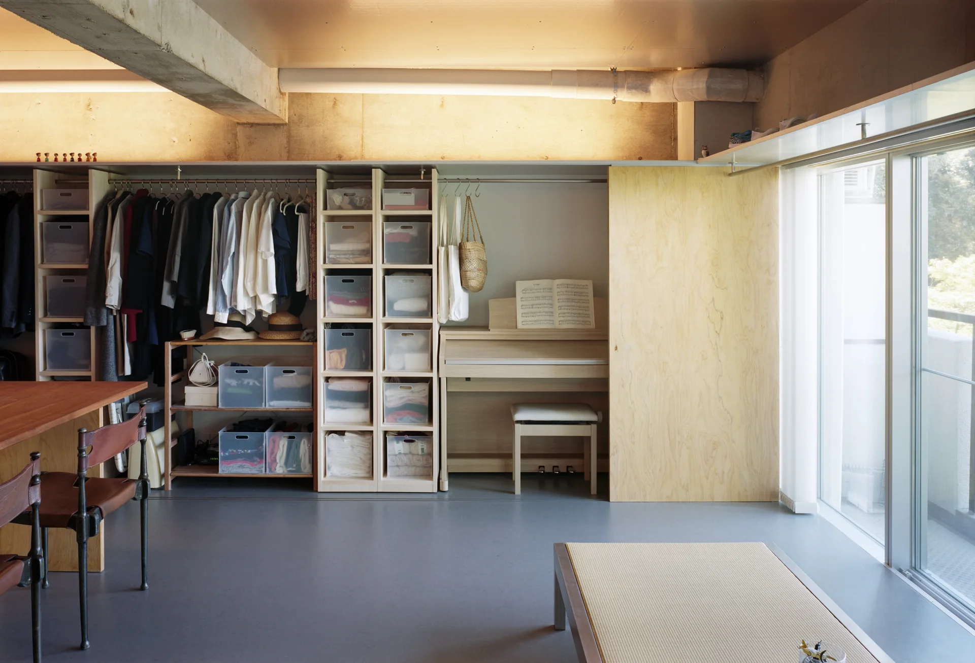 A minimalist wardrobe system with open shelving, hanging clothes, a digital piano, and a large sliding plywood door next to a window.