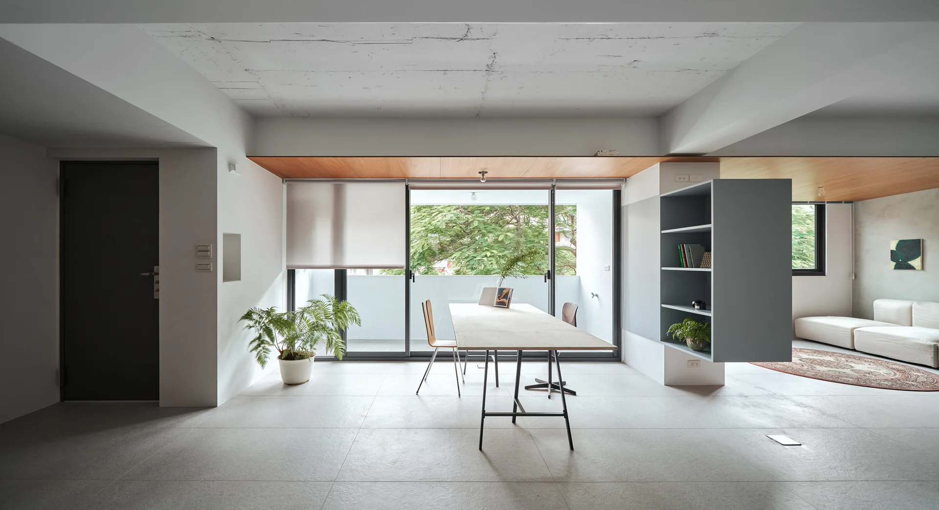 A minimalist open-plan dining room features a long wooden table, a gray shelving unit, sliding glass doors, and an exposed concrete ceiling.