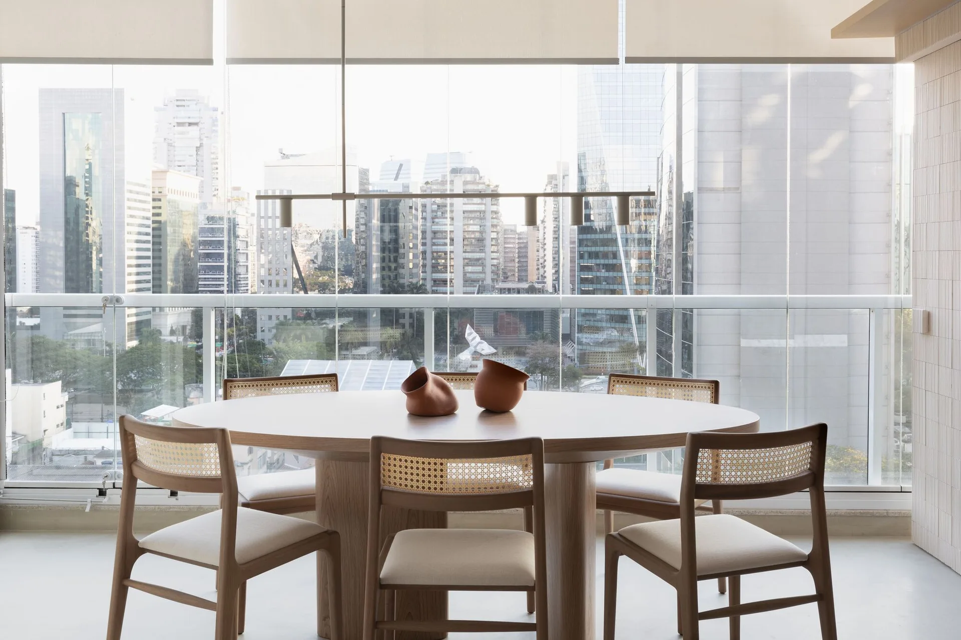 A light wood oval dining table and matching cane-backed chairs sit before a floor-to-ceiling glass window overlooking a city skyline.