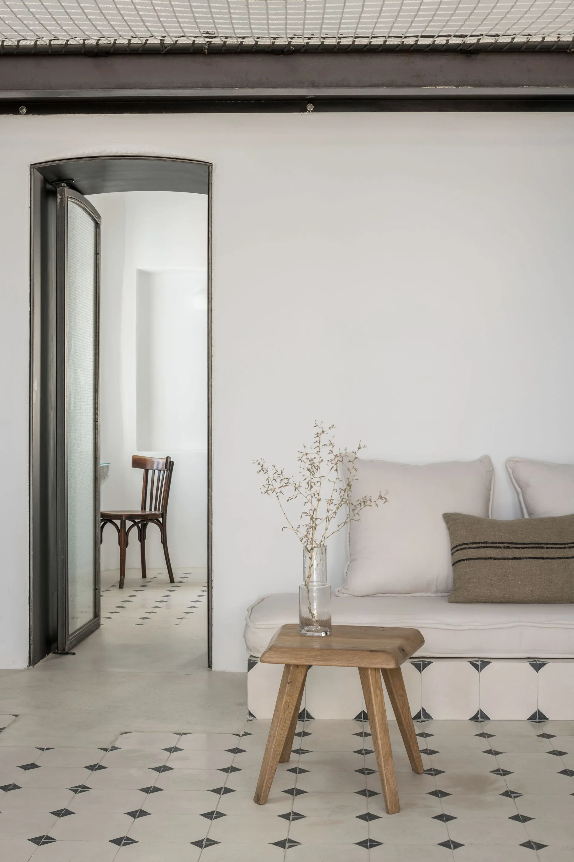 A bright living room features a built-in upholstered bench with a tiled base, a small wooden stool, and an arched doorway leading to a back room.