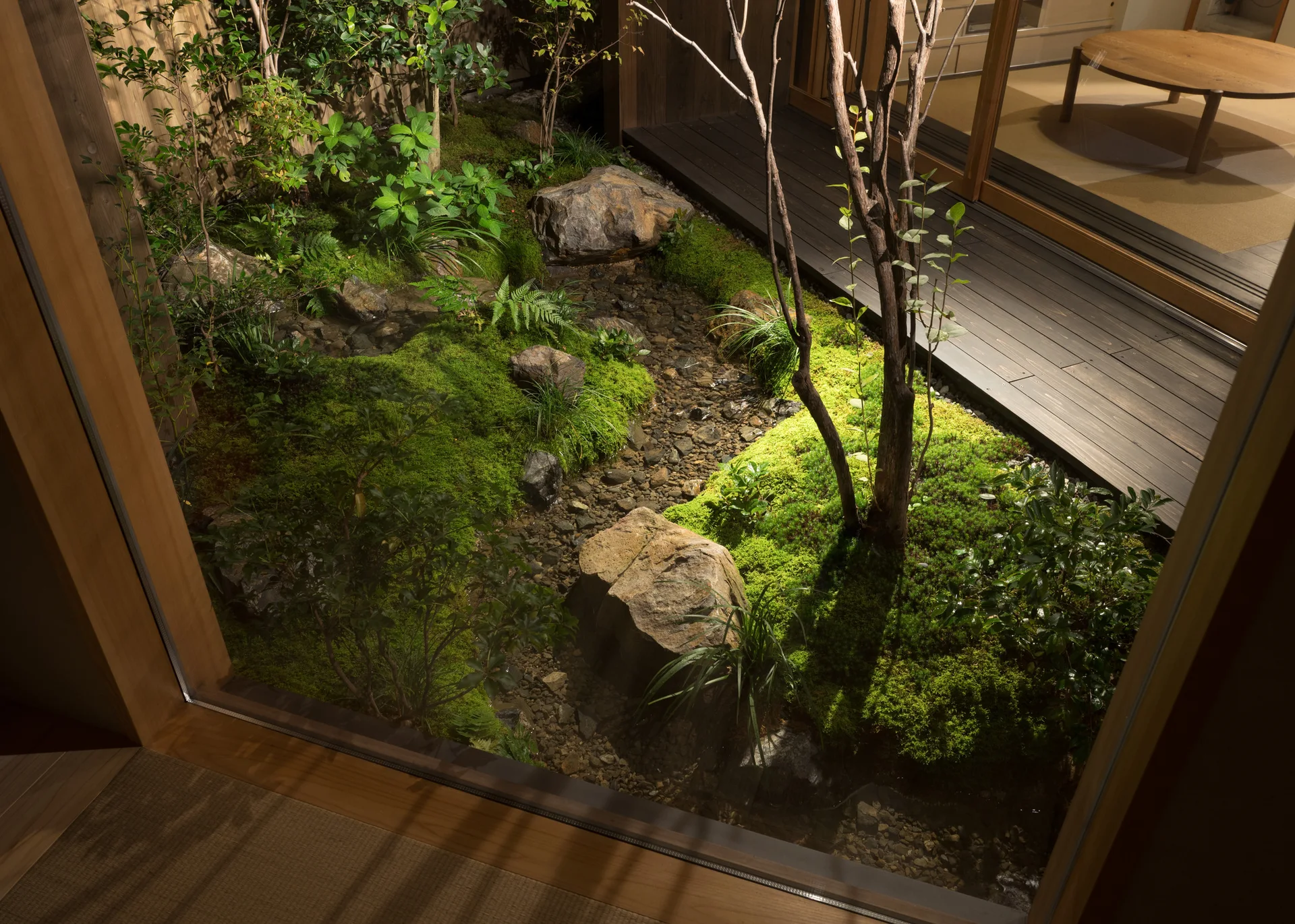 An indoor view looking through a large glass window onto a lush Japanese moss garden with natural boulders and a dark wood deck.