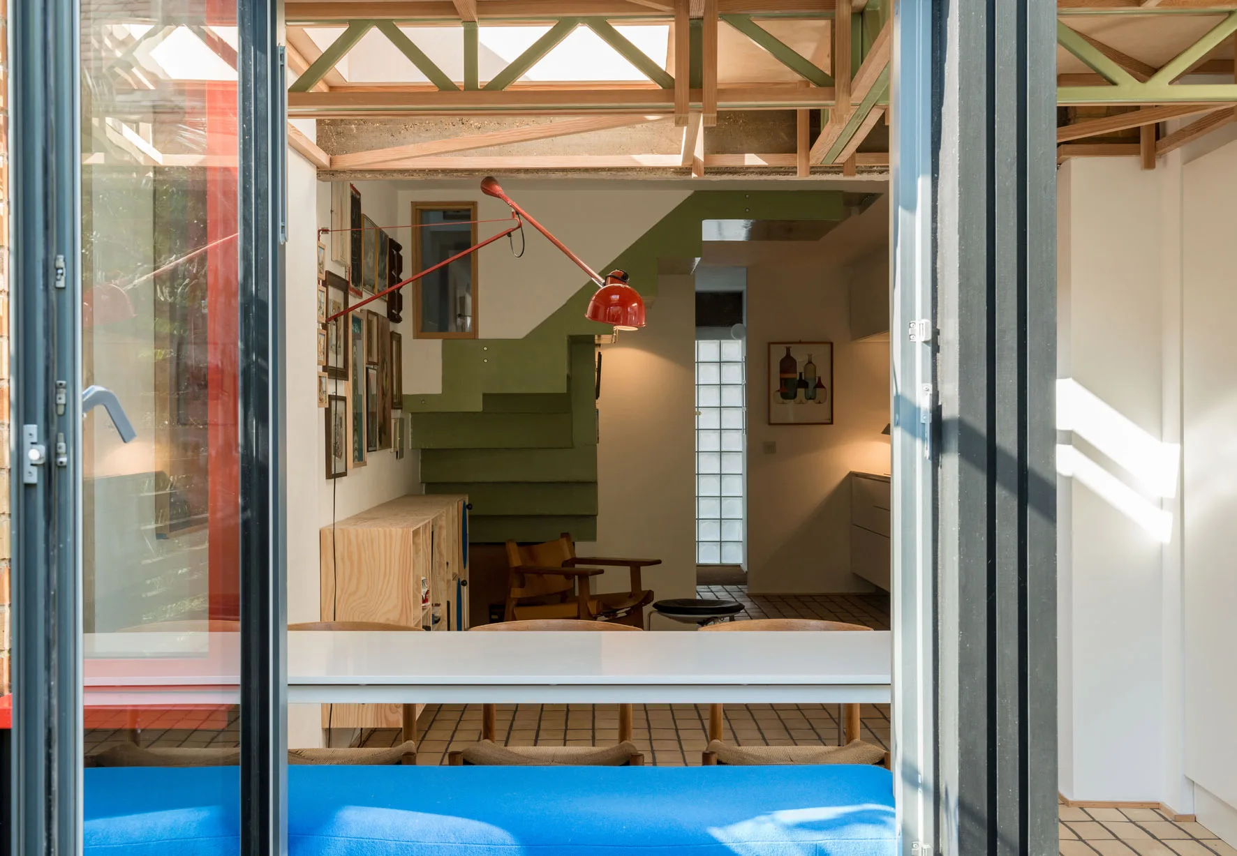 An interior dining area viewed through a glass door, featuring a white table, a green staircase, exposed wooden ceiling trusses, and a red wall lamp.
