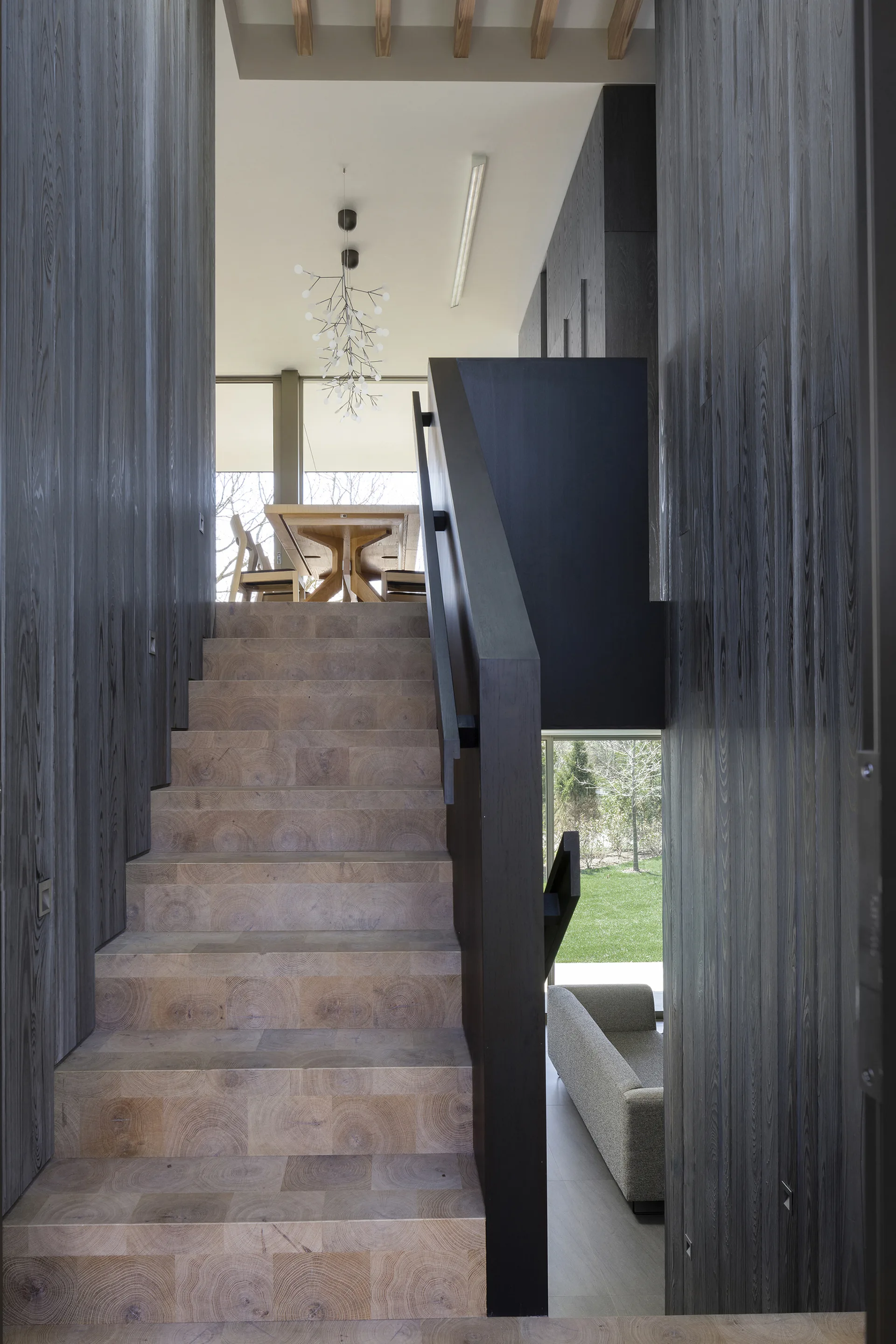 A modern interior staircase features light end-grain wood treads set between dark charcoal vertical wood paneled walls, leading up to a dining area.