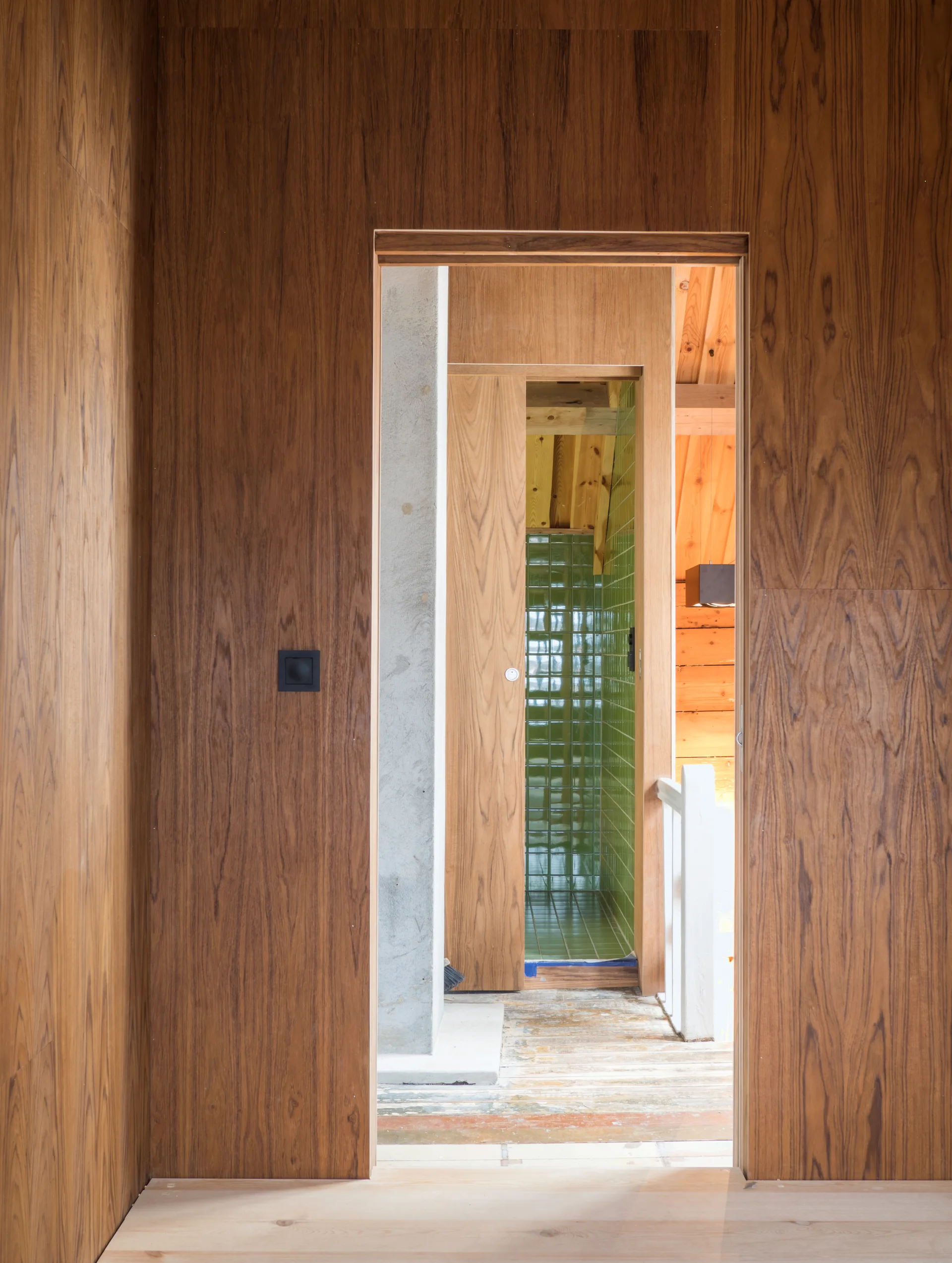 A view through a wood-grain paneled doorway showing a concrete pillar, a sliding wood door, and glossy green tiled walls.