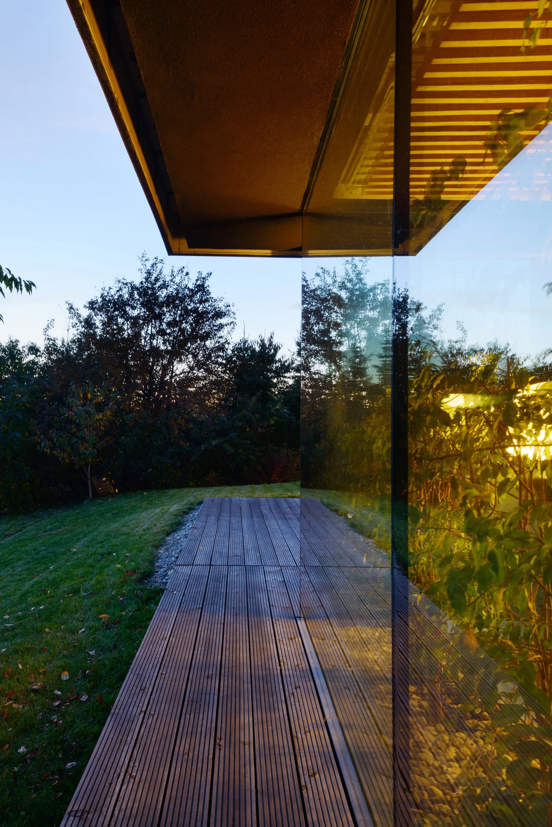 A modern wooden plank deck next to a reflective glass exterior wall showing reflections of trees and evening light.