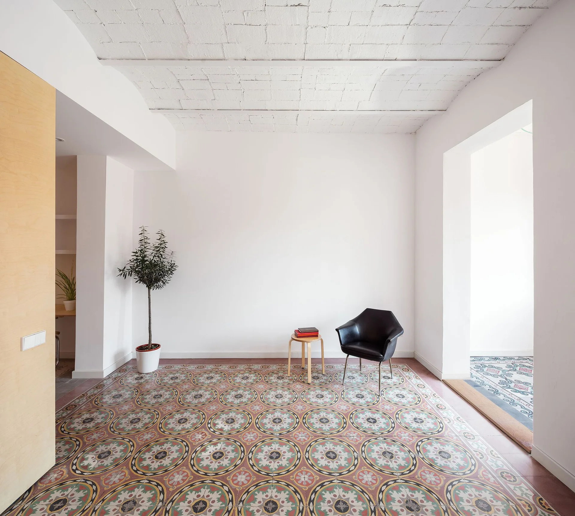 A minimalist white room with an ornate patterned tile floor, a black armchair, a wooden side table, and a potted tree.