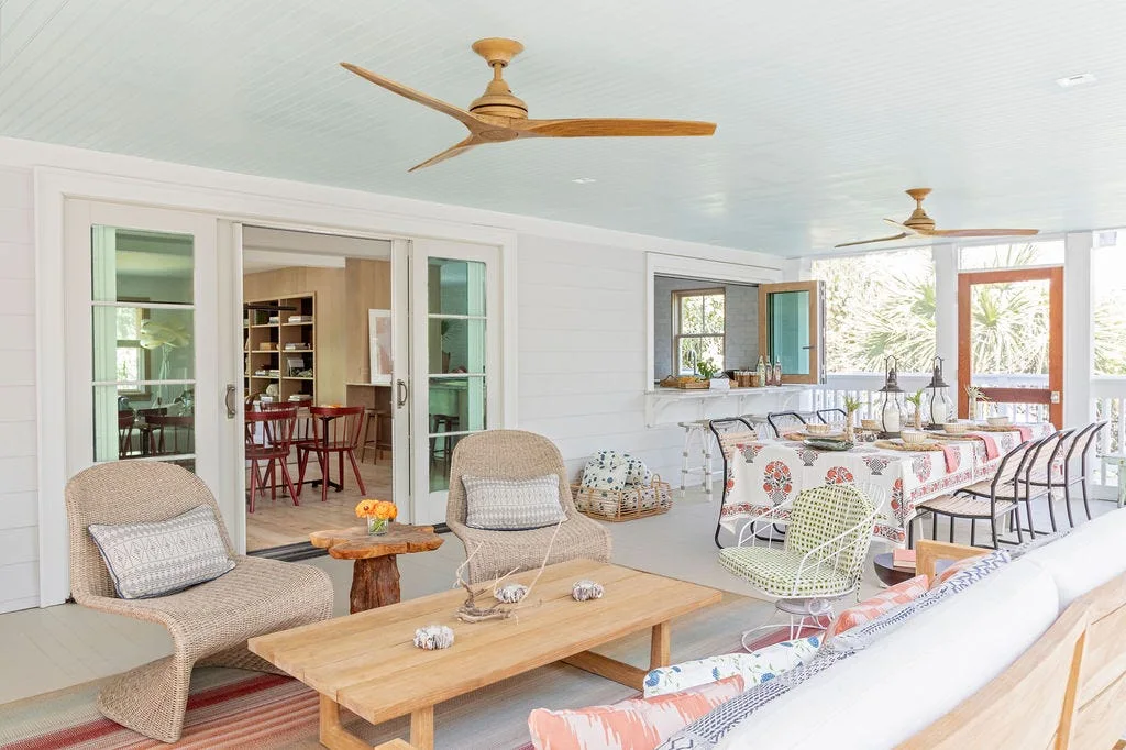 A spacious screened porch features a light blue wood-planked ceiling with two wooden fans, wicker lounge chairs, and a decorated dining table.