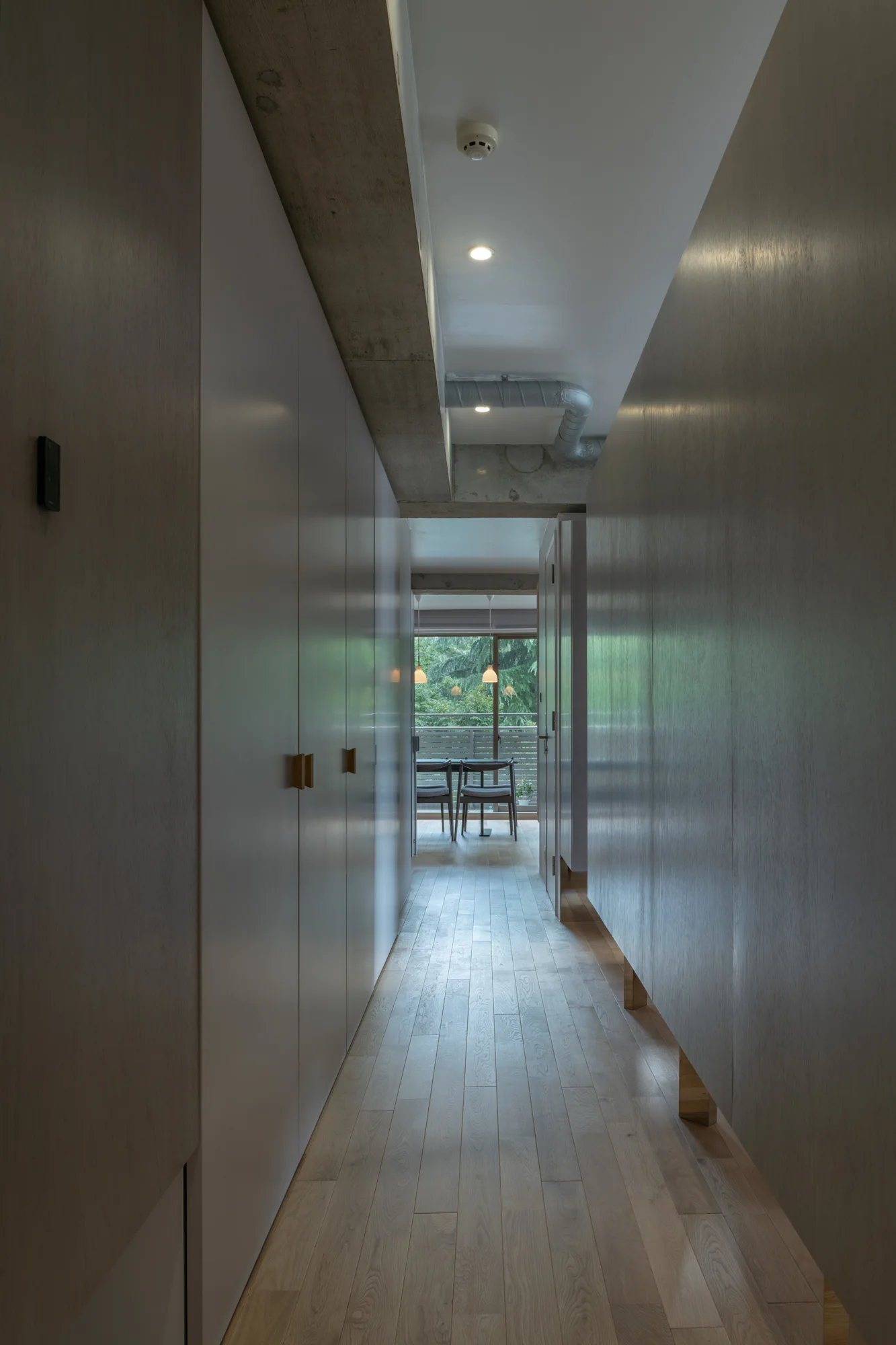 A narrow hallway with light wood cabinets on both sides and an exposed concrete ceiling beam leads to a bright dining room in the distance.