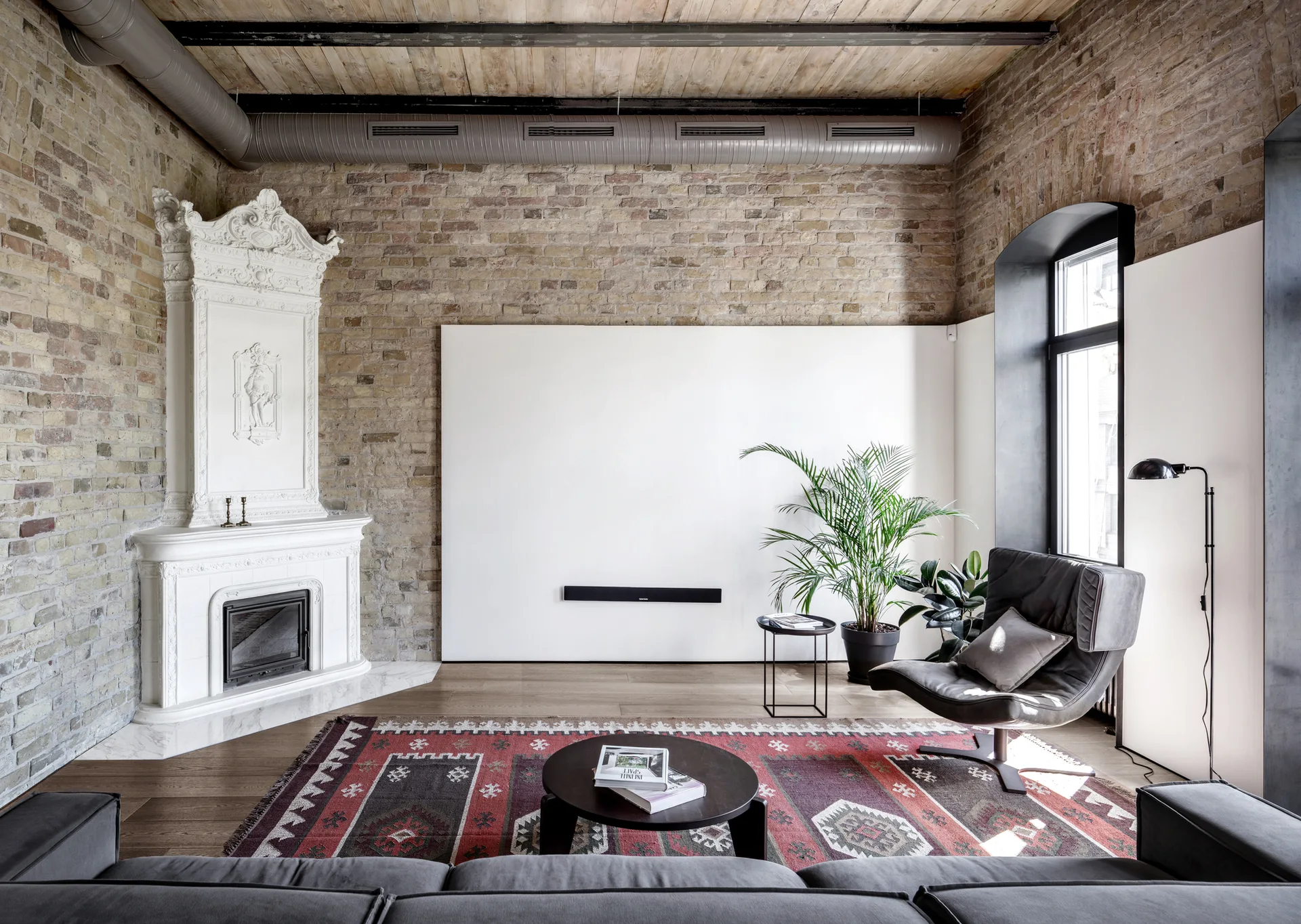 A loft living room features an ornate white fireplace, exposed brick walls, a traditional patterned rug, and a modern leather armchair.